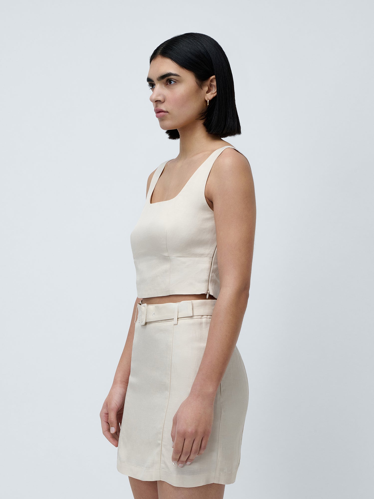 Eine Frau mit glattem schwarzem Haar trägt das schmal geschnittene „Majella“-Top von LeGer by Lena Gercke und einen dazu passenden Rock mit hoher Taille und Gürtel. Sie steht seitlich vor einem schlichten hellgrauen Hintergrund und präsentiert damit einen frischen Sommerlook.