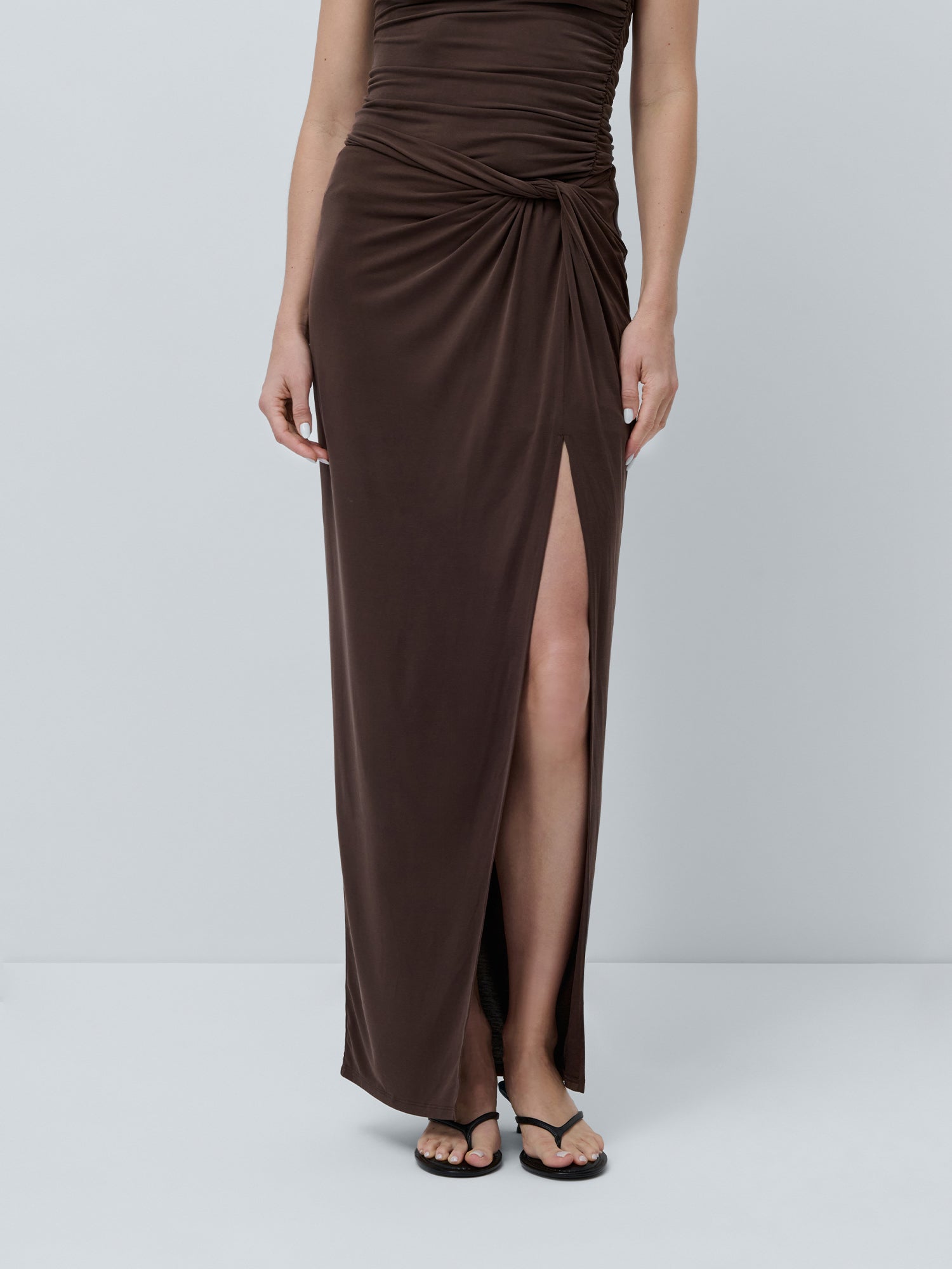 Eine Person trägt den Perle Skirt von LeGer by Lena Gercke, einen dunkelbraunen, knöchellangen Rock mit hoher Taille, asymmetrischem Design, hohem Schlitz und Raffung an der Taille. Dazu passen schwarze Sandalen vor einem schlichten, hellen Hintergrund.