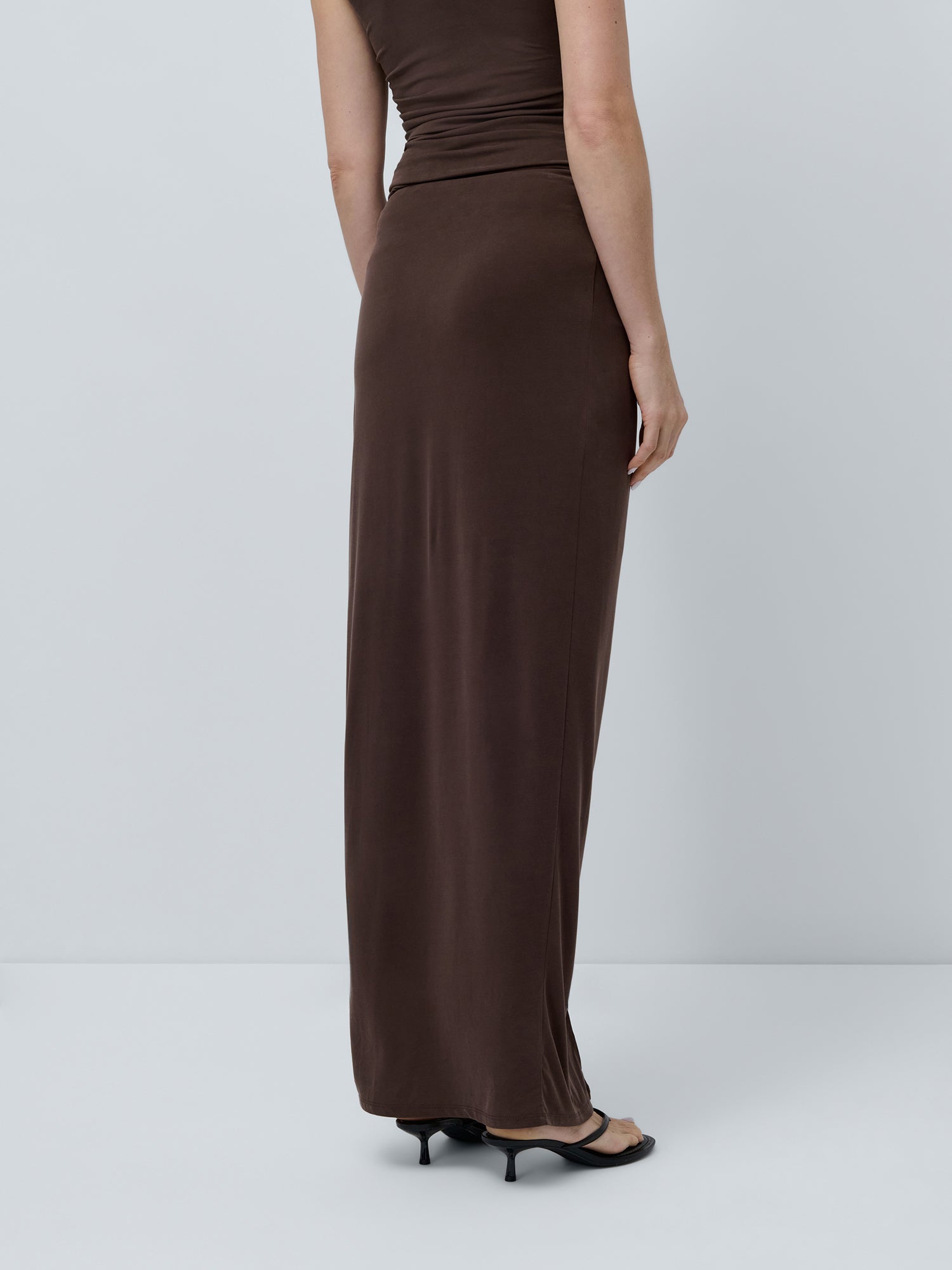 Eine Person trägt den Perle Rock von LeGer by Lena Gercke als knöchellangen Rock mit asymmetrischem Design, kombiniert mit einem braunen, ärmellosen Top und schwarzen Heels. Der Look ist seitlich vor hellem Hintergrund zu sehen.