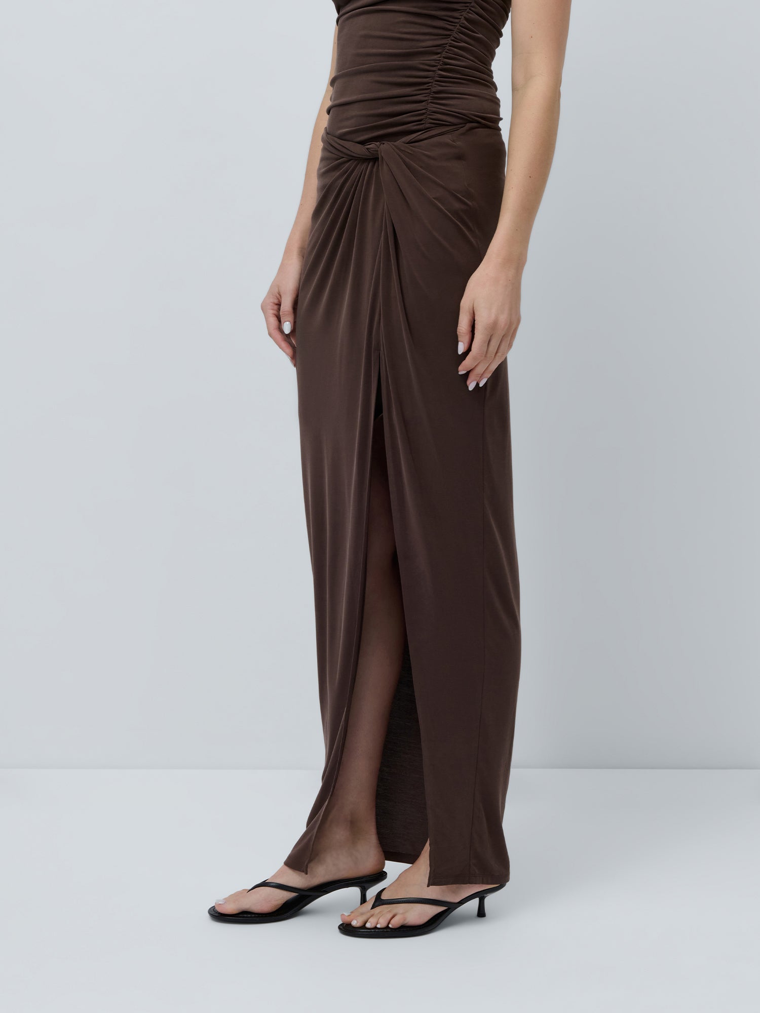 Eine Person präsentiert den Perle-Rock von LeGer by Lena Gercke – ärmellos, dunkelbraun mit gerafften und asymmetrischen Details sowie einem hohen Schlitz vorne – kombiniert mit schwarzen Sandalen mit Absatz vor einem schlichten hellgrauen Hintergrund.