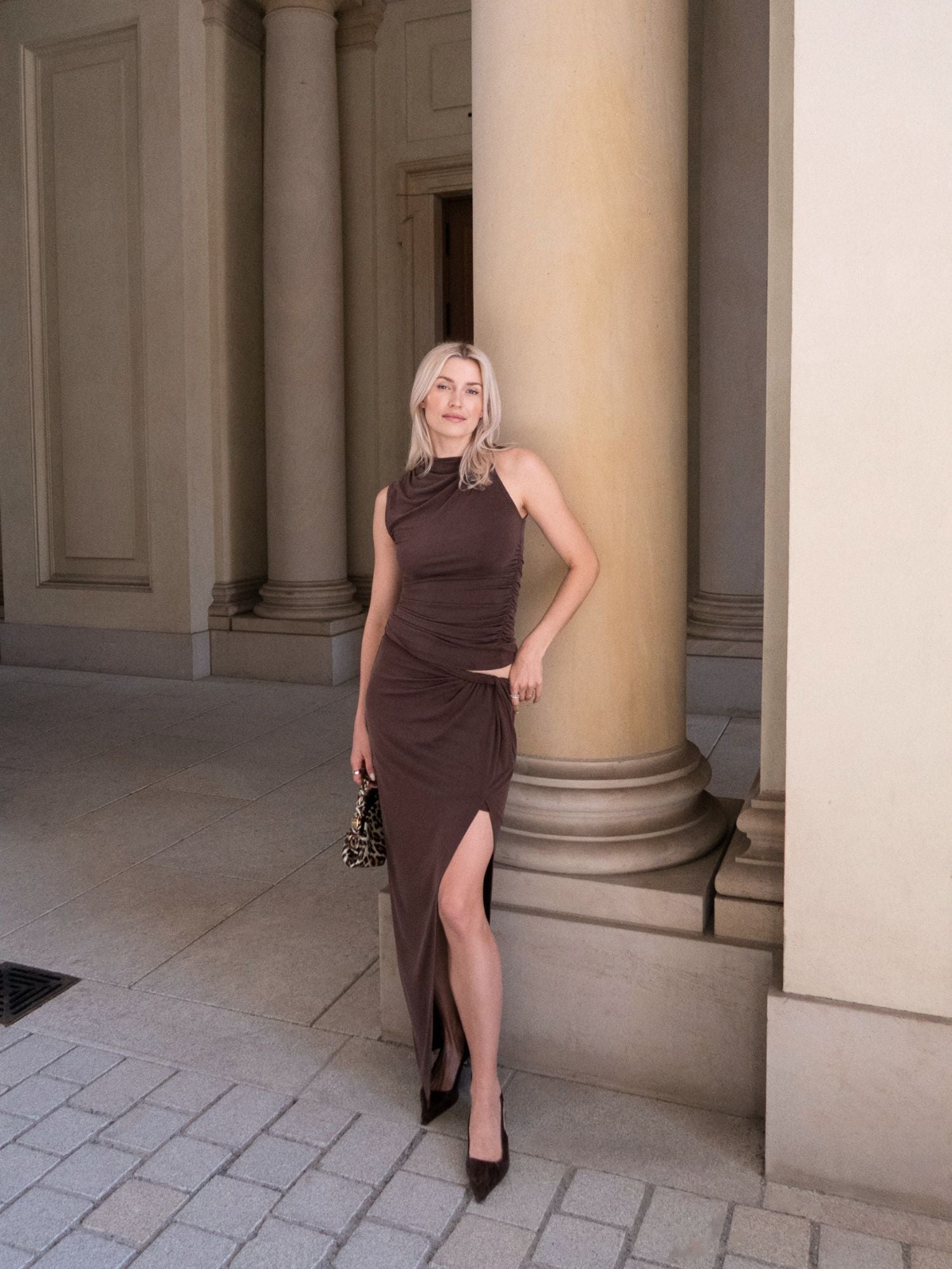 Eine Frau präsentiert den Rock „Perle“ von LeGer by Lena Gercke, der sich durch eine hohe Taille und einen oberschenkellangen Schlitz auszeichnet, kombiniert mit schwarzen Pumps und einer kleinen, gemusterten Handtasche. Sie steht vor einer klassischen Säule inmitten von Steinböden und hohen Säulen.