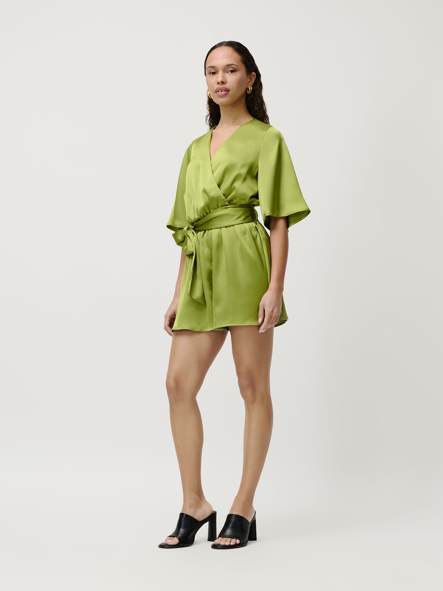 Eine Frau mit langem Haar und neutralem Gesichtsausdruck präsentiert mühelose Mode im Alexis Overall von LeGer by Lena Gercke. Er besteht aus einem hellgrünen Mini-Wickeldesign aus Satin mit Taillenband, weiten Ärmeln und wird mit schwarzen Sandalen mit offenen Zehen und Absatz kombiniert.