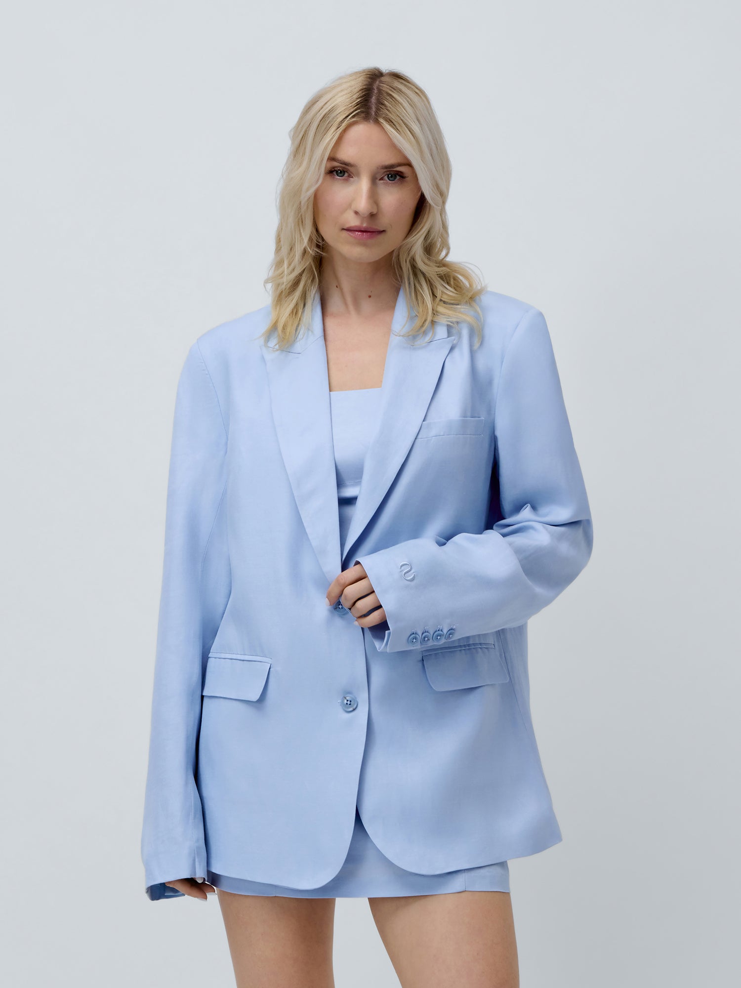 Eine blonde Frau trägt den hellblauen Oversize-Blazer „Majella“ mit langen Ärmeln von LeGer by Lena Gercke und einen dazu passenden Rock. Sie steht vor einem schlichten hellgrauen Hintergrund und blickt mit ausdruckslosem Gesichtsausdruck in die Kamera.