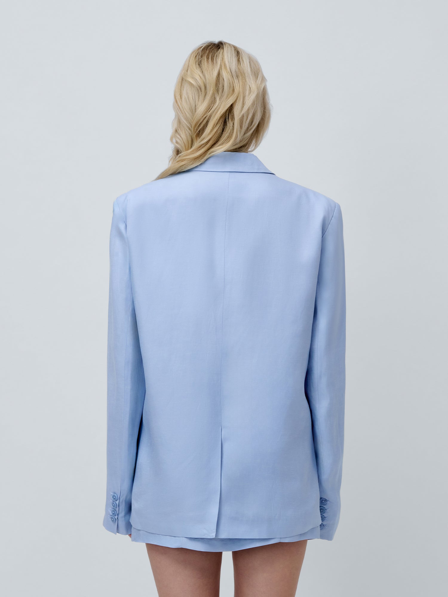 Eine Person mit langen blonden Haaren blickt von der Kamera weg, trägt den hellblauen Majella Blazer von LeGer by Lena Gercke mit auffälligen Schulterpolstern und ohne sichtbare Hose und posiert vor einem schlichten hellgrauen Hintergrund.