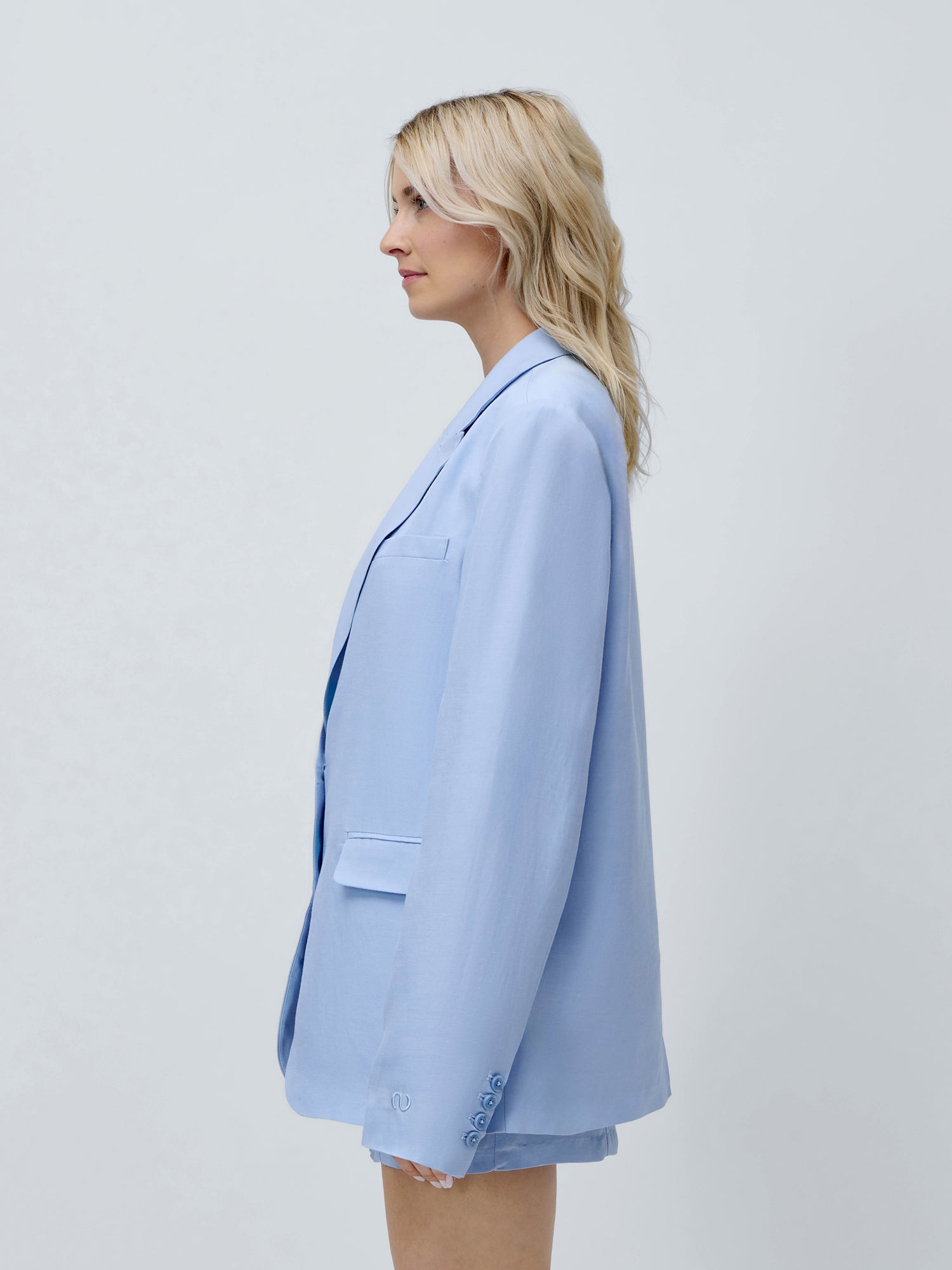 Eine Frau mit welligem blondem Haar steht im Profil vor einem schlichten hellgrauen Hintergrund und trägt einen hellblauen „Majella“-Blazer von LeGer by Lena Gercke sowie dazu passende Shorts.