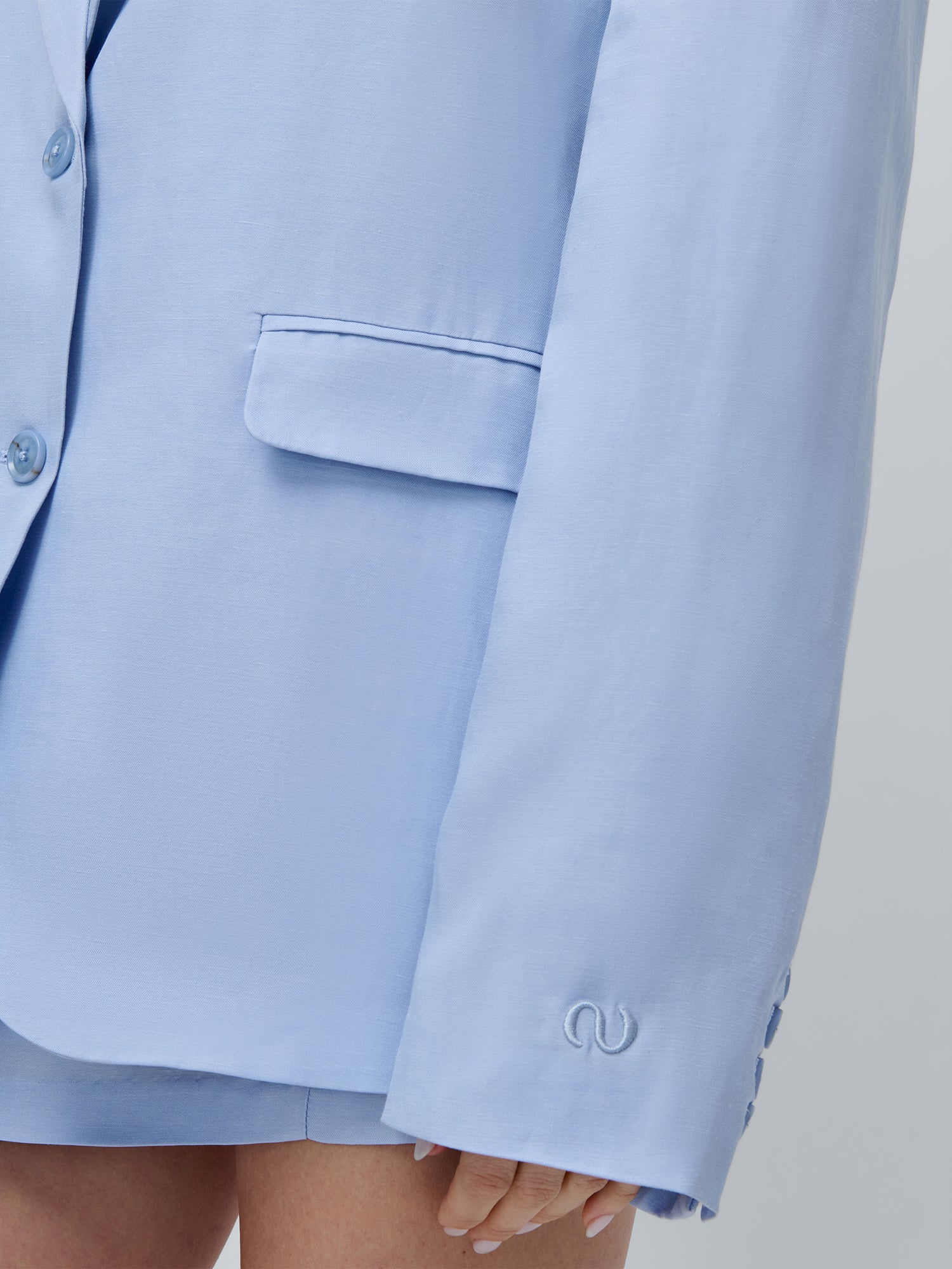 Eine Nahaufnahme einer Person, die den hellblauen Majella Blazer von LeGer by Lena Gercke mit passenden Shorts trägt. Der Blazer hat eine Vordertasche, dezente Stickereien an der Manschette, klassische Revers und die Hand der Person ist teilweise sichtbar.