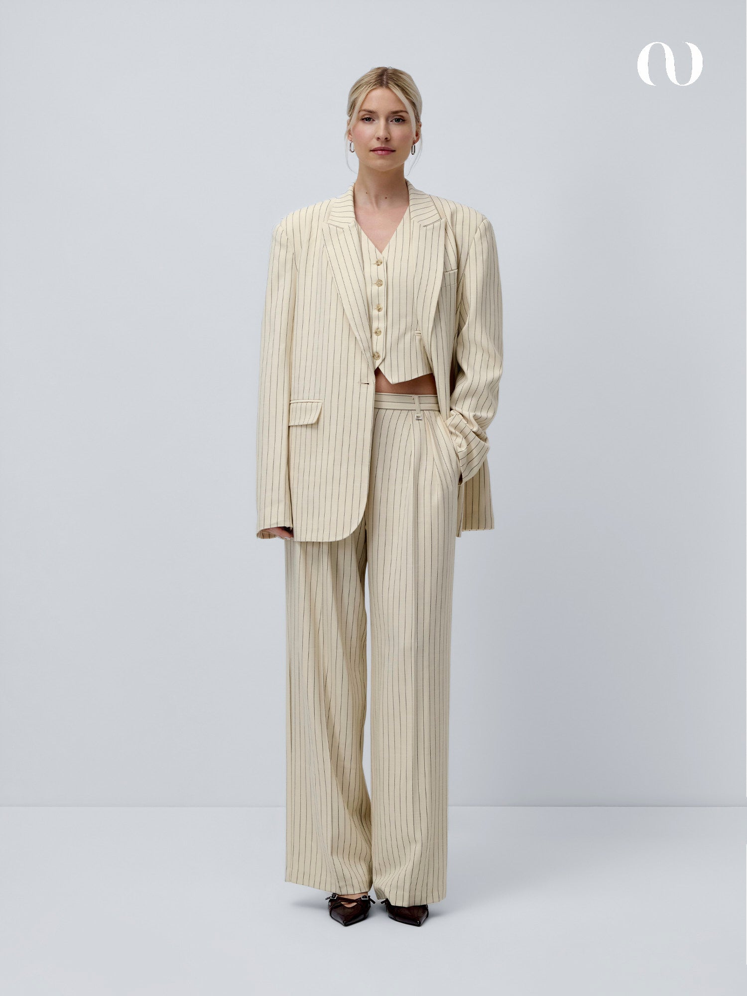 Eine Frau steht vor einem schlichten weißen Hintergrund und trägt den Elaine Blazer von LeGer by Lena Gercke - einen übergroßen, cremefarbenen Nadelstreifenanzug mit extralangen Ärmeln, einer Hose mit weitem Bein, einer geknöpften Weste und schwarzen spitzen Schuhen. Ihr Haar ist zurückgebunden.