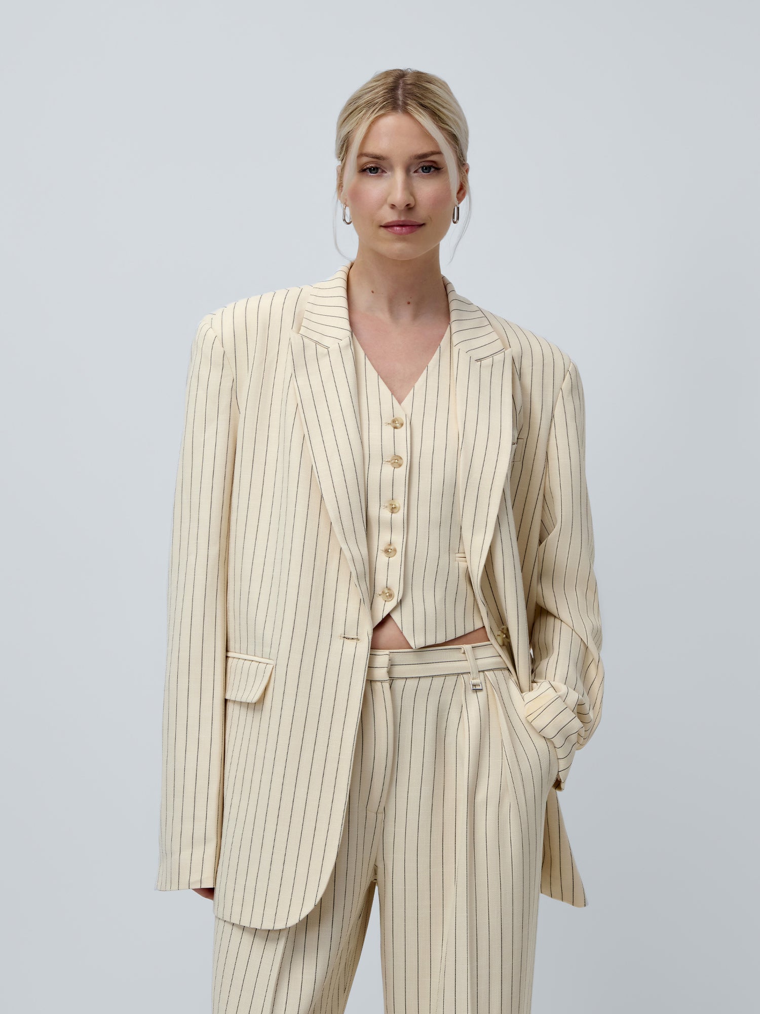 Vor einem hellgrauen Hintergrund steht eine Frau selbstbewusst und trägt einen cremefarbenen Blazer mit Nadelstreifen, eine Weste und eine Hose aus der Kollektion „Elaine“ von LeGer by Lena Gercke. Ihr blondes Haar ist zu einem tief sitzenden Dutt gestylt.
