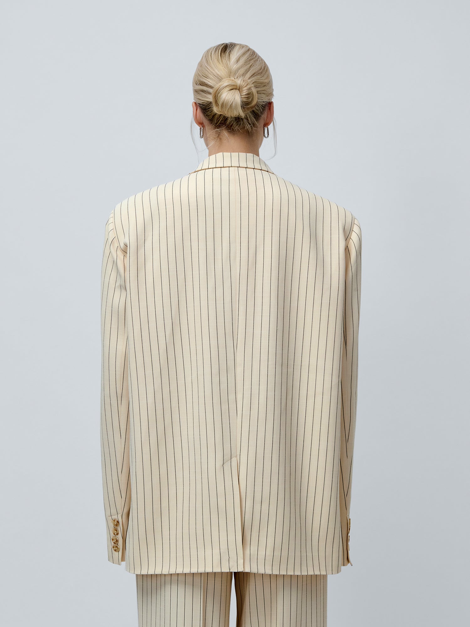 Eine Person mit blonden Haaren, die zu einem tief sitzenden Dutt zusammengebunden sind, ist von hinten zu sehen. Sie trägt den cremefarbenen Blazer „Elaine“ von LeGer by Lena Gercke, der sich durch feine vertikale schwarze Streifen auf einem einfarbigen hellgrauen Hintergrund auszeichnet.