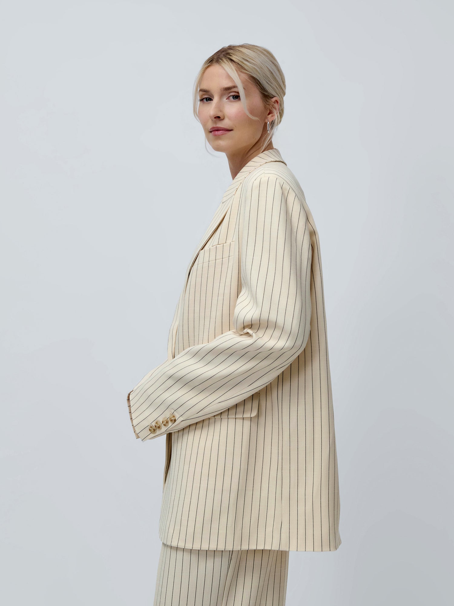 Eine Frau mit blonden Haaren, die zu einem tief sitzenden Dutt zusammengebunden sind, steht im Profil vor einem hellgrauen Hintergrund. Sie trägt den Blazer „Elaine“ von LeGer by Lena Gercke, der in einem übergroßen, cremefarbenen Nadelstreifen-Design gestaltet ist.