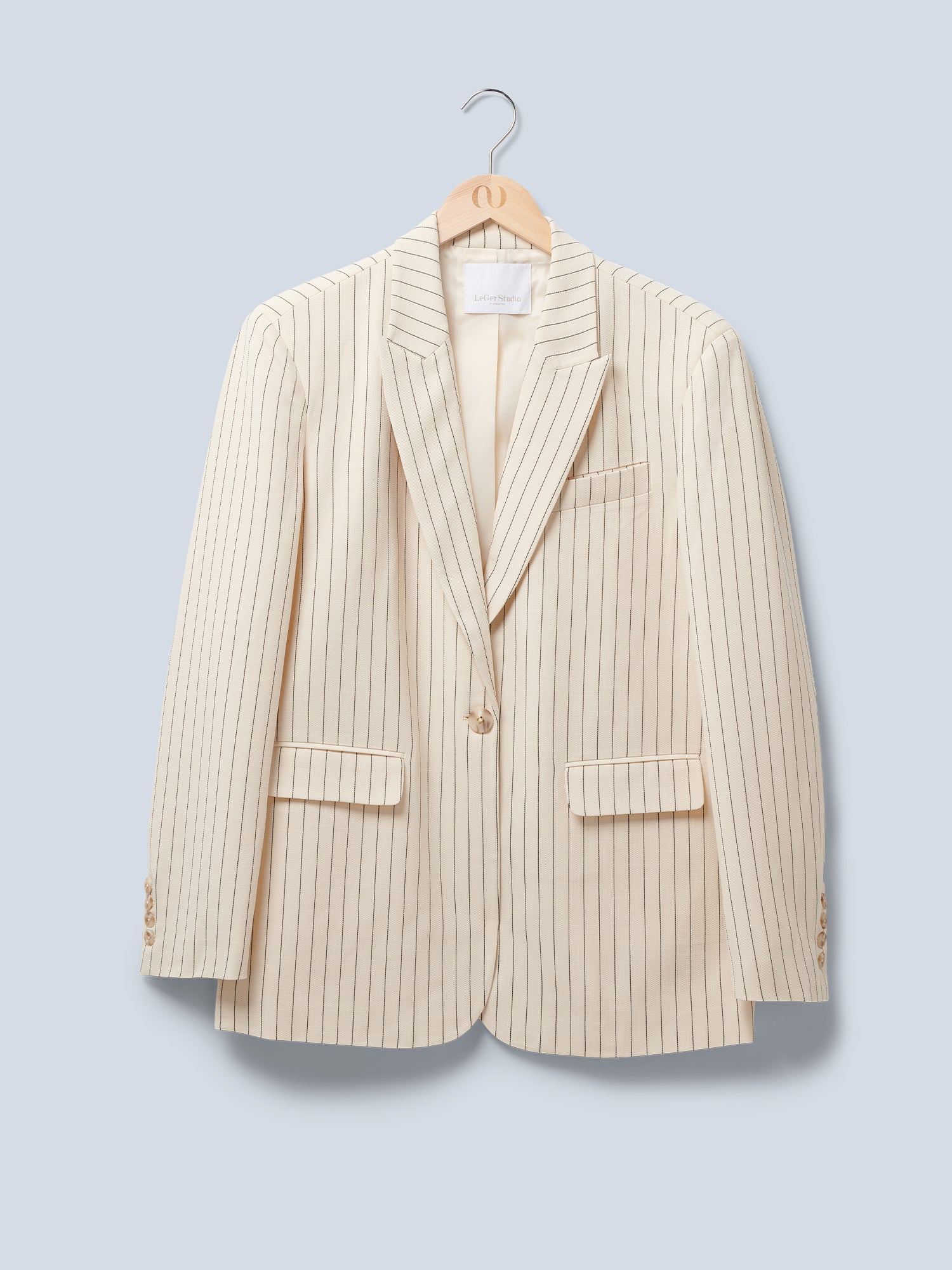 Der cremefarbene Blazer „Elaine“ von LeGer by Lena Gercke besticht durch feine schwarze Nadelstreifen, ein fallendes Revers, zwei Pattentaschen vorne und einen Ein-Knopf-Verschluss. Er wird in Oversize-Passform auf einem Holzkleiderbügel vor einem hellgrauen Hintergrund präsentiert.