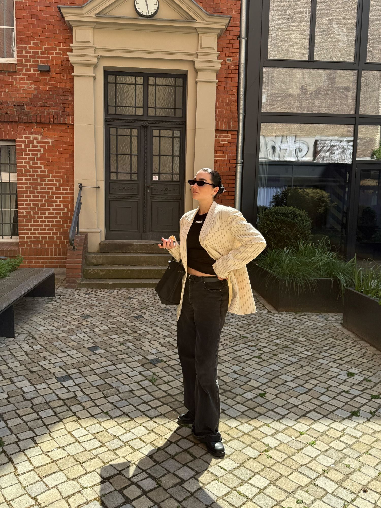 Eine Frau mit Sonnenbrille, einem gestreiften, übergroßen „Elaine“-Blazer von LeGer by Lena Gercke, einem schwarzen Oberteil und einer dunklen Jeans steht auf einem gepflasterten Innenhof, hält ihr Handy in der Hand und blickt neben einem Backsteingebäude mit einer Uhr über der Tür nach oben.