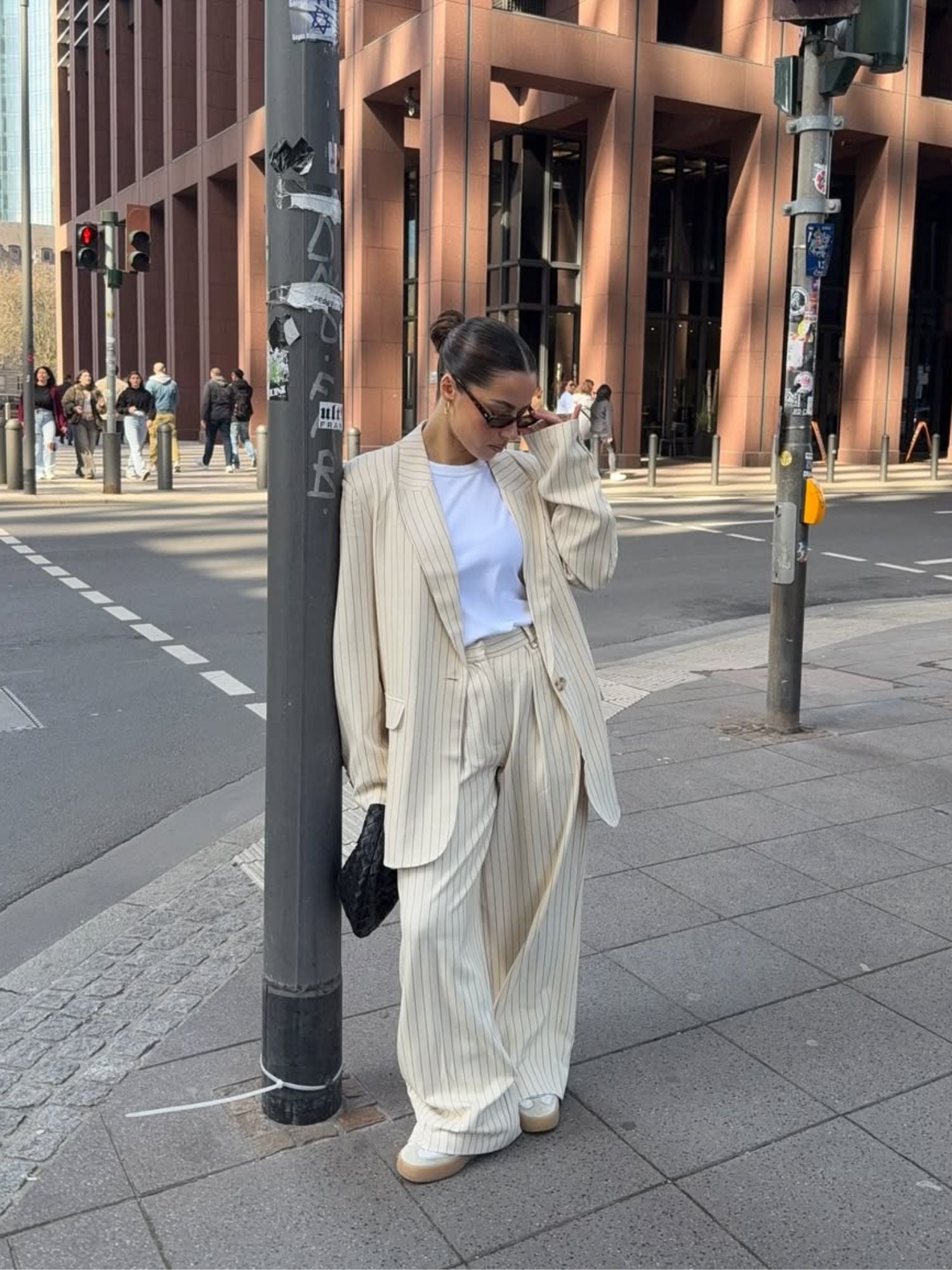 Eine Person steht an einer Straßenecke in der Stadt und trägt einen übergroßen, beigen „Elaine“-Blazer von LeGer by Lena Gercke, dazu ein weißes T-Shirt und eine Sonnenbrille. Sie lehnt an einem Pfosten, während Menschen zwischen hohen Gebäuden die Straße überqueren.
