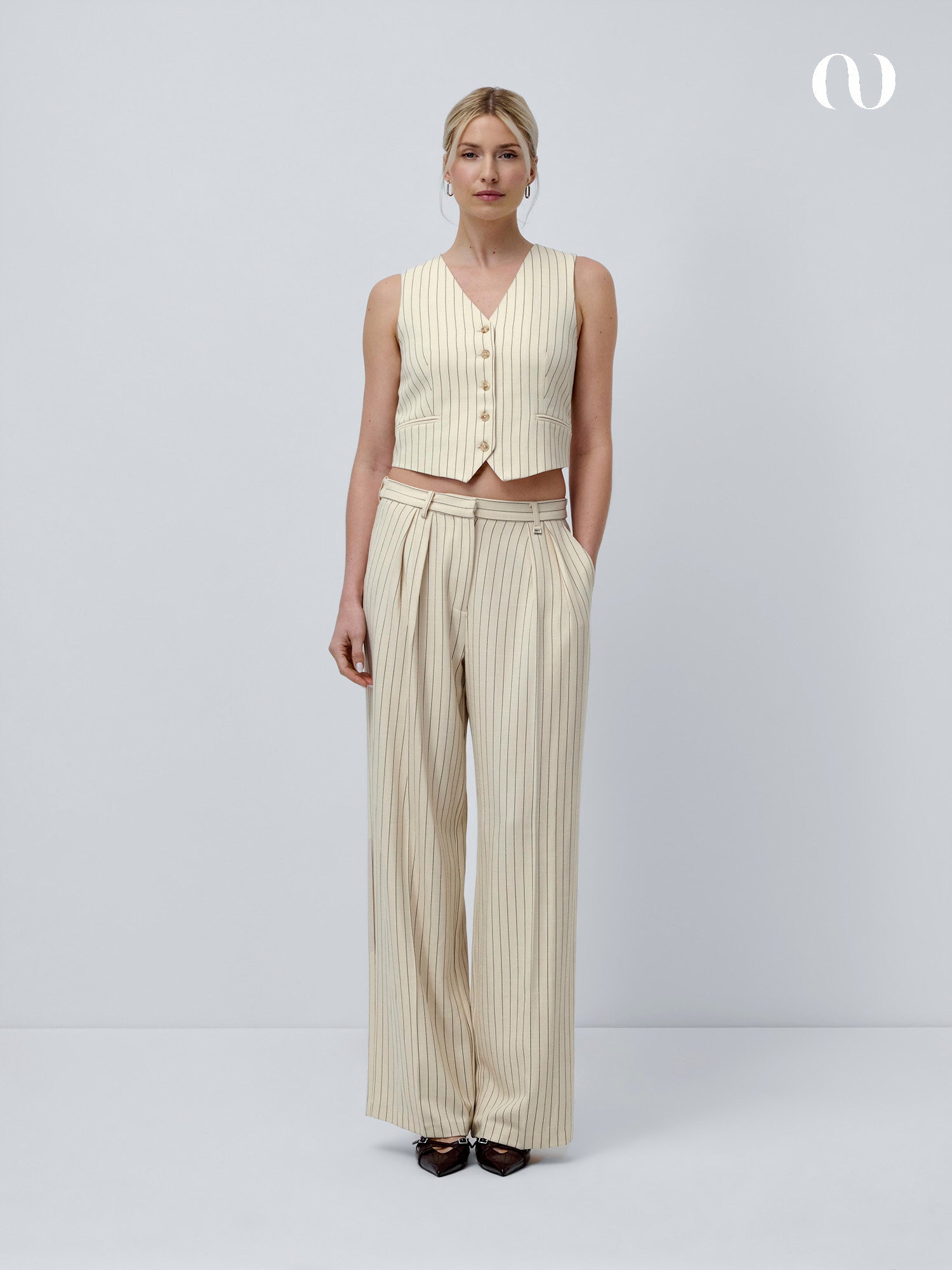 Eine Frau steht vor einem unifarbenen, hellgrauen Hintergrund und trägt die Elaine Vest von LeGer by Lena Gercke mit einer dazu passenden, hoch taillierten Hose mit weitem Bein und verstellbarem Bund. Sie hat die Hände in den Taschen und trägt dunkle Schuhe.