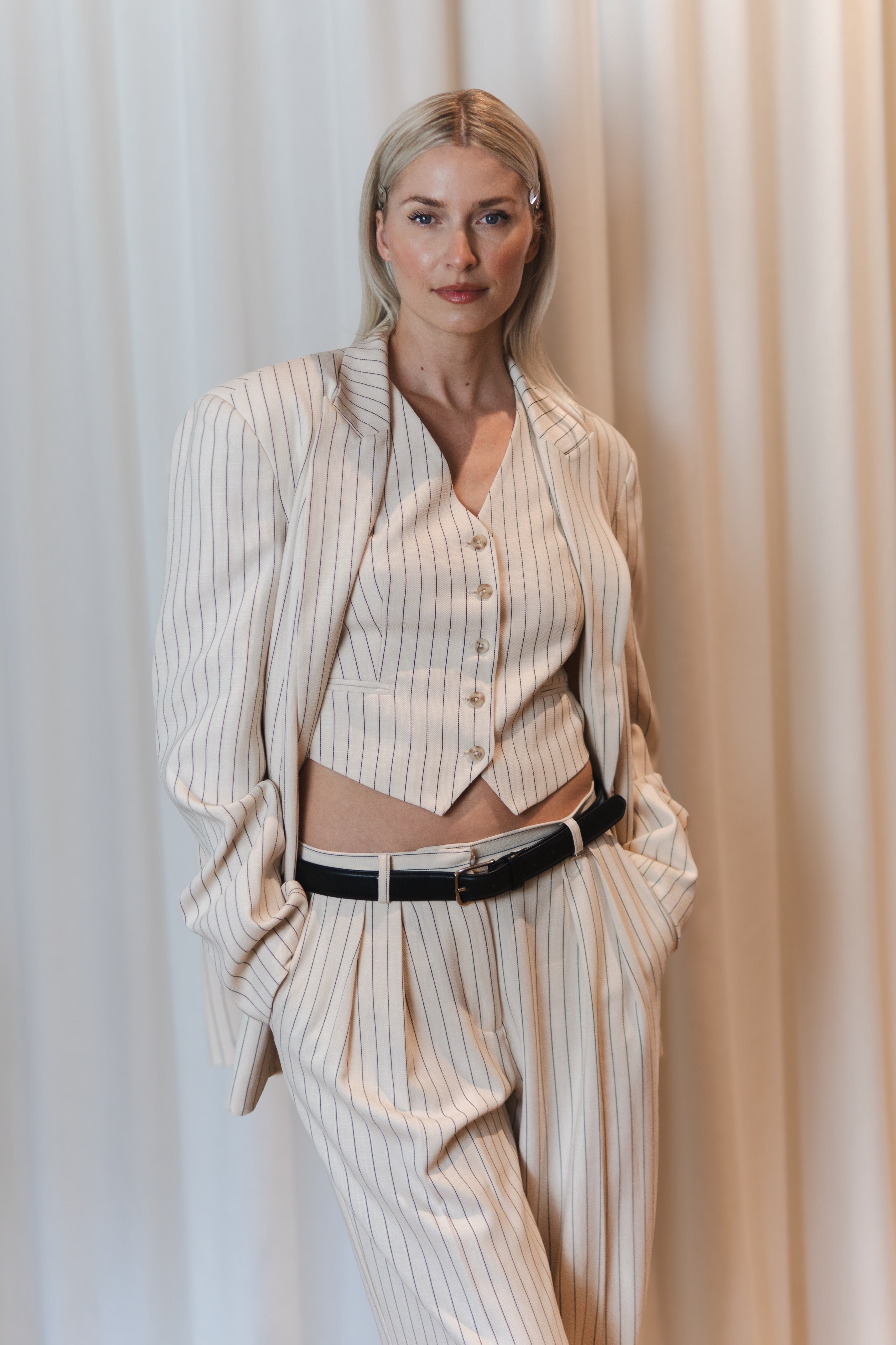 Eine Frau mit glattem blondem Haar modelliert die Elaine Vest von LeGer by Lena Gercke, eine cremefarbene ärmellose Weste mit Nadelstreifen und V-Ausschnitt und einer lockeren Hose. Sie posiert selbstbewusst mit einer Hand in der Tasche vor einem sanft drapierten Vorhang.
