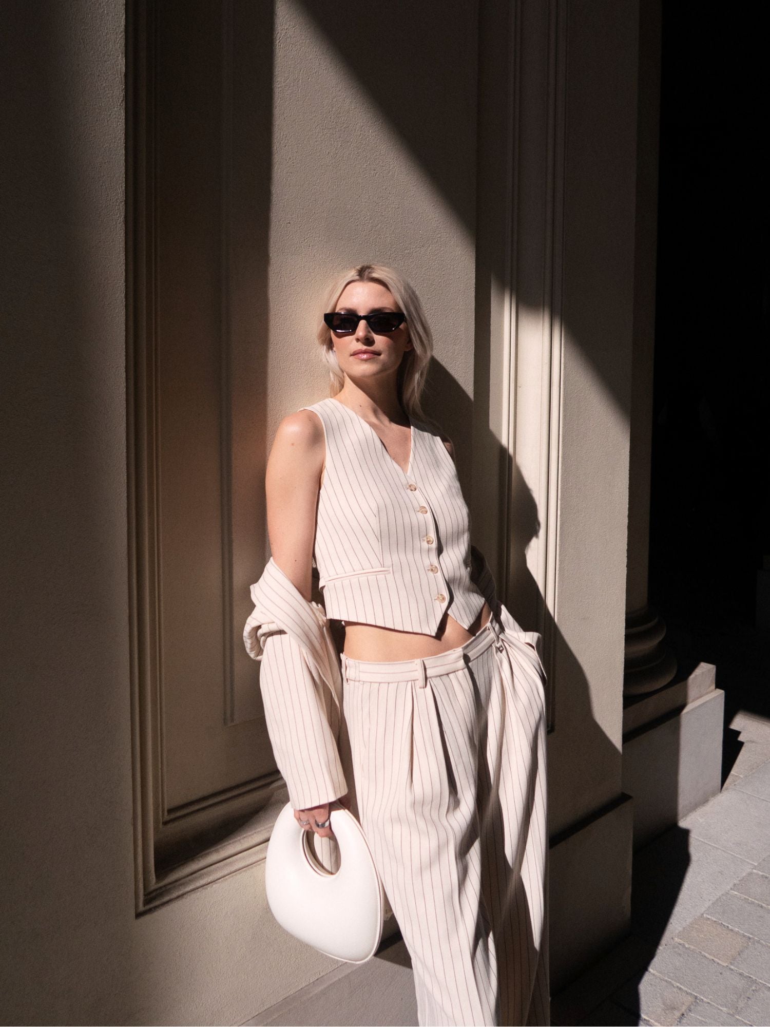 Eine Frau mit Sonnenbrille posiert im Sonnenlicht. Sie trägt die Elaine Vest von LeGer by Lena Gercke mit einer passenden Hose mit weitem Bein, eine über den Arm gehangene Jacke und eine weiße Handtasche. Sie steht vor einer hohen, strukturierten Wand.