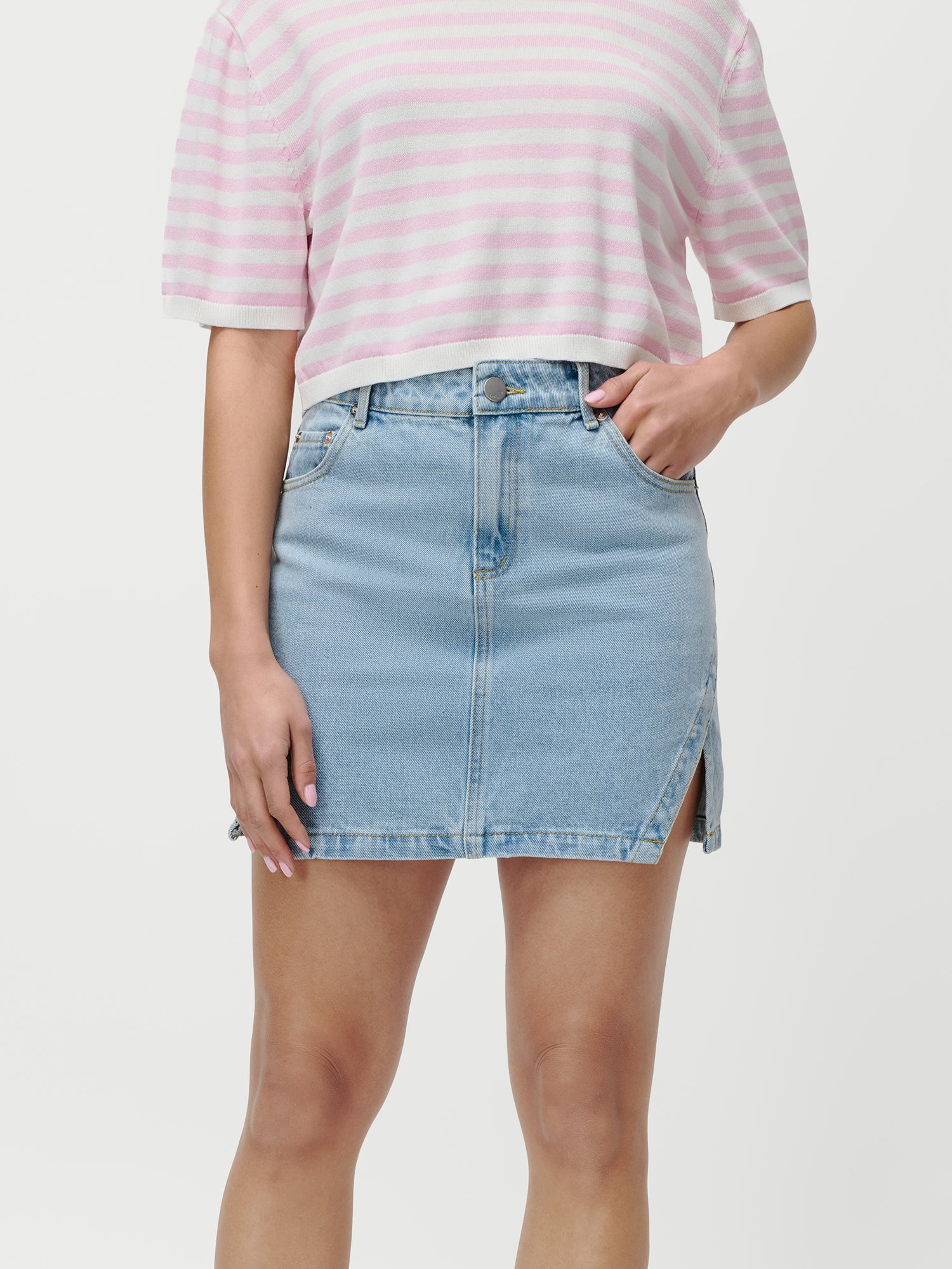 Eine Person, die ein hellrosa und weiß gestreiftes kurzärmeliges Oberteil und den Daphne Rock von LeGer by Lena Gercke aus hellblauem Jeansstoff mit einem Schlitz an der Vorderseite trägt, steht vor einem schlichten weißen Hintergrund.