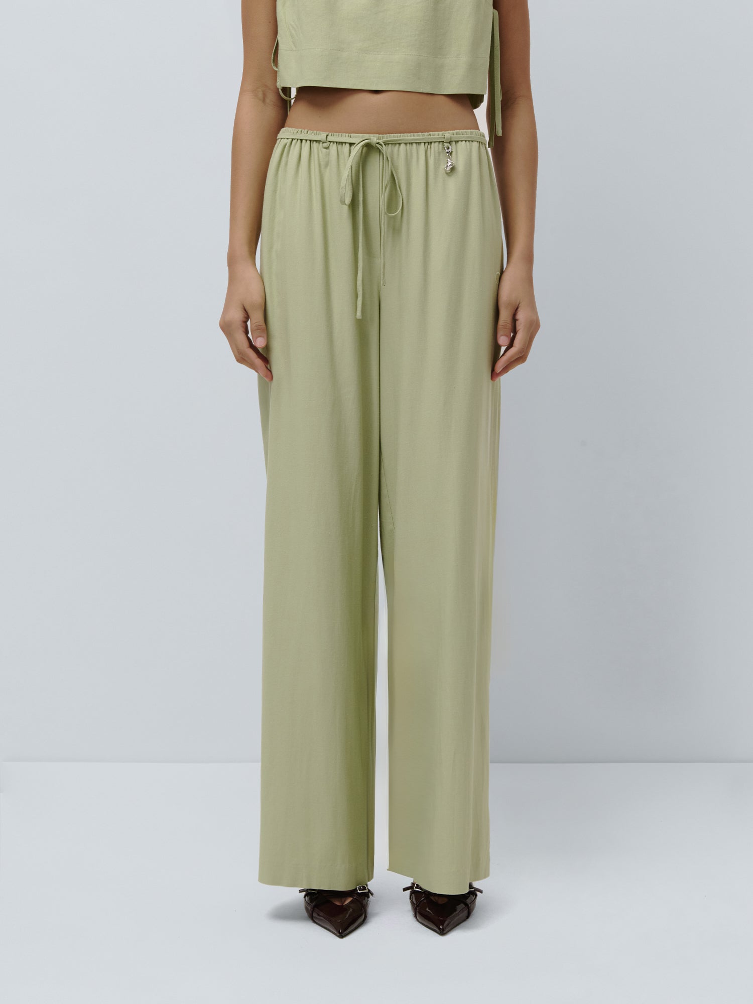 Eine Person trägt eine hellgrüne, locker sitzende „Emelie“-Hose von LeGer by Lena Gercke sowie ein dazu passendes kurzes Oberteil und steht vor einem schlichten hellgrauen Hintergrund. Nur die untere Körperhälfte ist zu sehen.