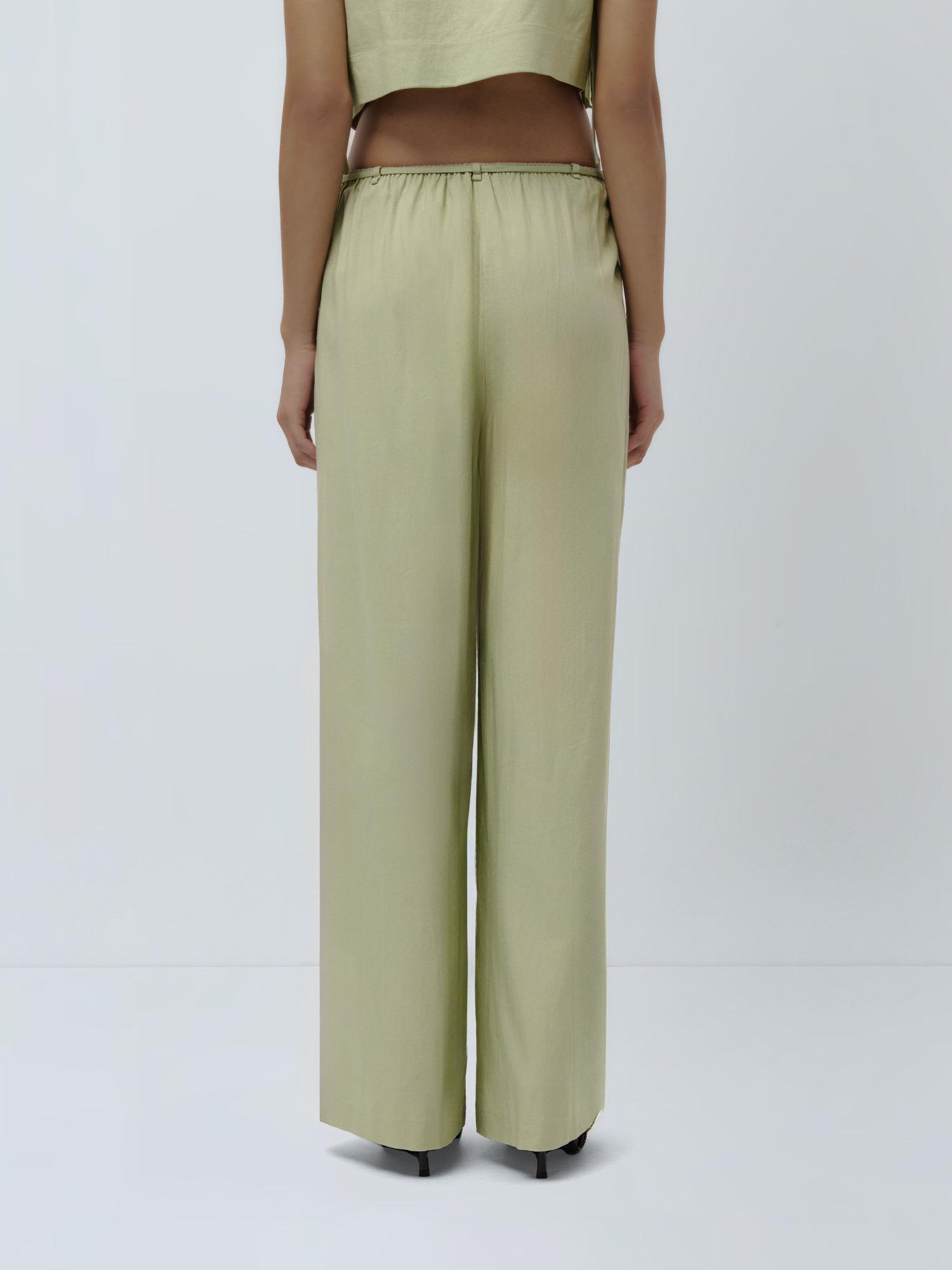 Eine Person steht mit dem Rücken zur Kamera und trägt die hoch taillierte, locker sitzende Hose „Emelie“ von LeGer by Lena Gercke aus hellgrünem Satin sowie ein dazu passendes kurzes Oberteil vor einem schlichten weißen Hintergrund.