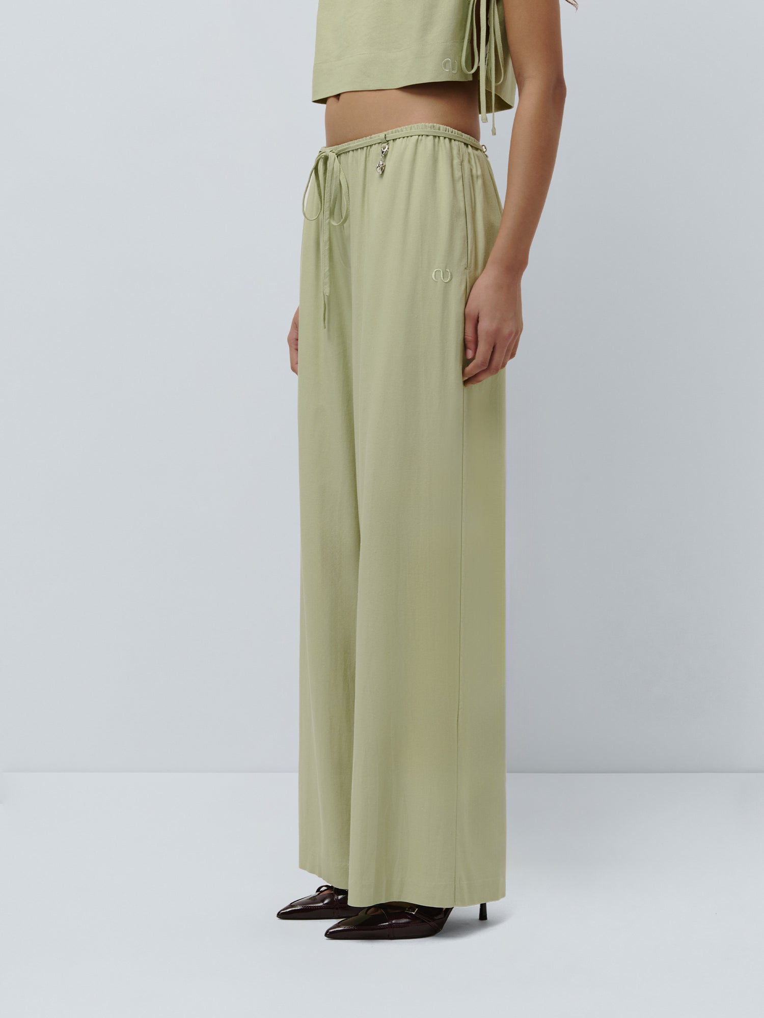 Eine Person steht im Profil und trägt die Hose „Emelie“ von LeGer by Lena Gercke in Hellgrün mit Paperbag-Taille und weitem Bein, kombiniert mit einem passenden Crop-Top mit seitlichen Bändern und spitzen dunkelbraunen Schuhen, vor einem schlichten hellen Hintergrund.