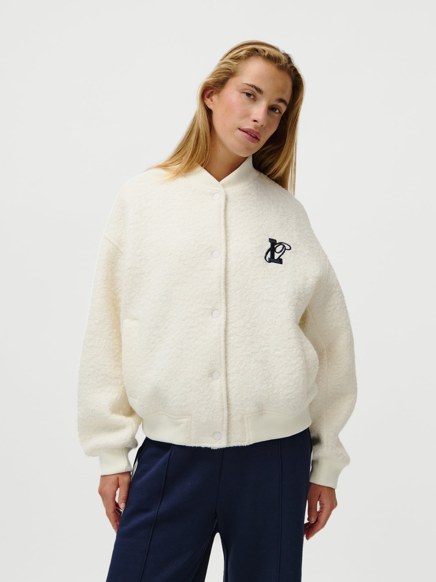 Eine Frau modelliert die LeGer x Oh April - Blanca Jacket von LeGer by Lena Gercke, ein cremefarbenes, langärmeliges Fleece mit Knopfleiste und dunklem Logo auf der linken Brust, gepaart mit einer marineblauen Hose, vor einem schlichten weißen Hintergrund.