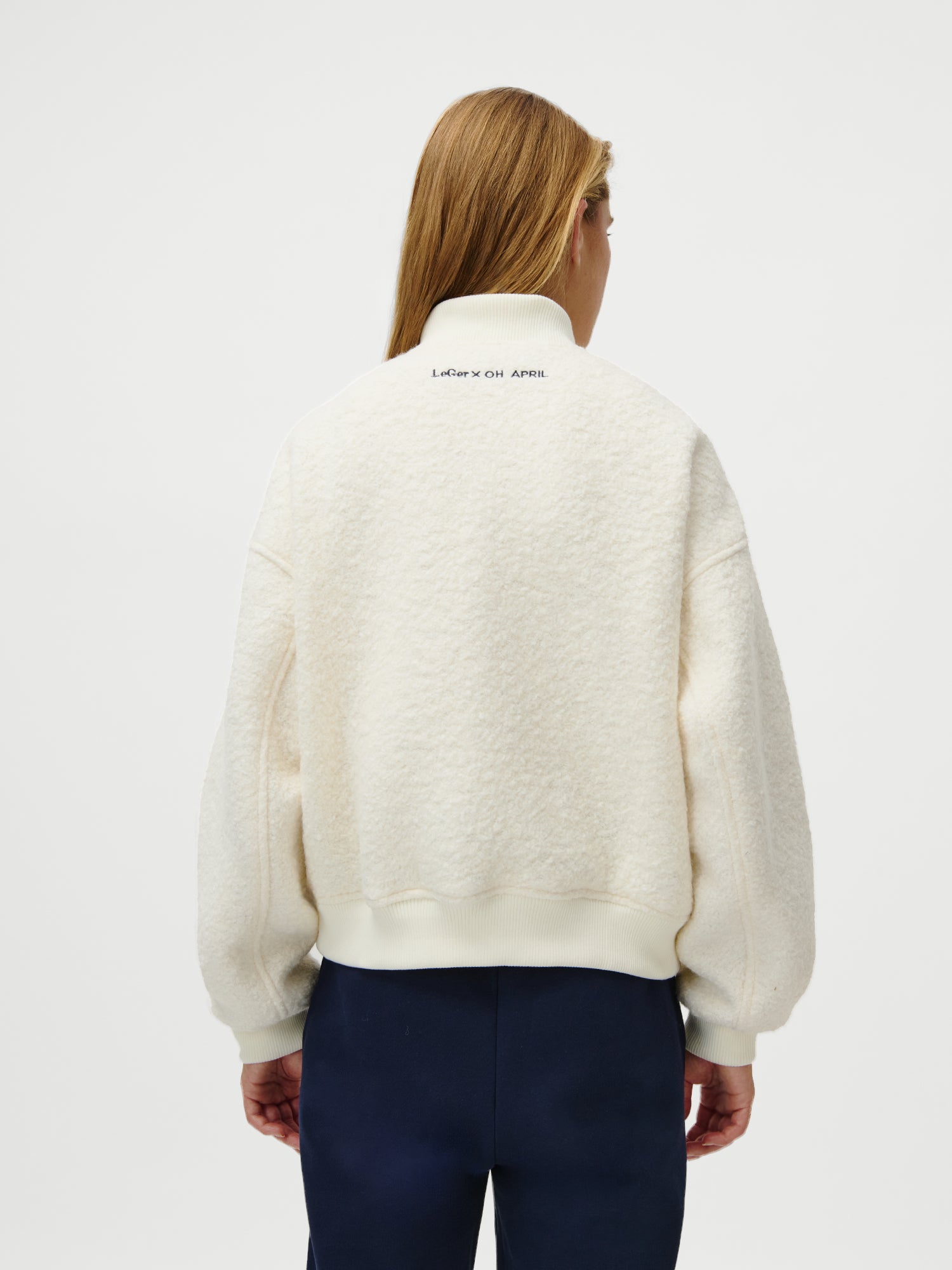 Eine Person mit langen, hellbraunen Haaren trägt die LeGer x Oh April - Blanca Jacket by LeGer by Lena Gercke, eine cremefarbene Jacke in normaler Länge mit geripptem Bund und Kragen, die einen kleinen Text im Nackenbereich aufweist. Sie wird mit einer dunkelblauen Hose kombiniert.