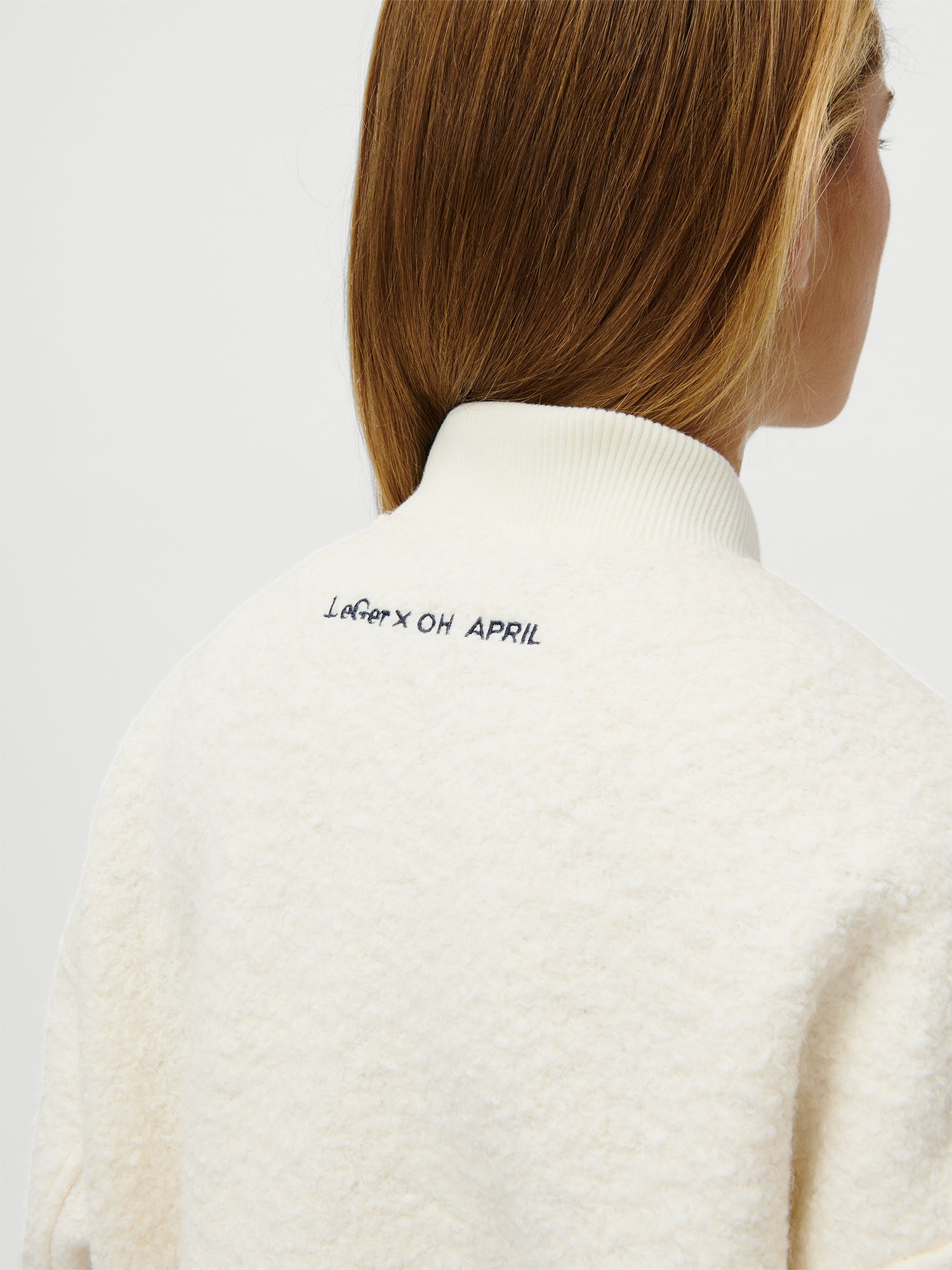 Abgebildet ist eine Person mit hellbraunem Haar von hinten, die die LeGer x Oh April - Blanca Jacket von LeGer by Lena Gercke trägt. Auf dem oberen Rücken der weißen, langärmeligen Jacke mit Rippkragen ist in schwarzer Farbe "LeGer x Oh April" eingestickt.