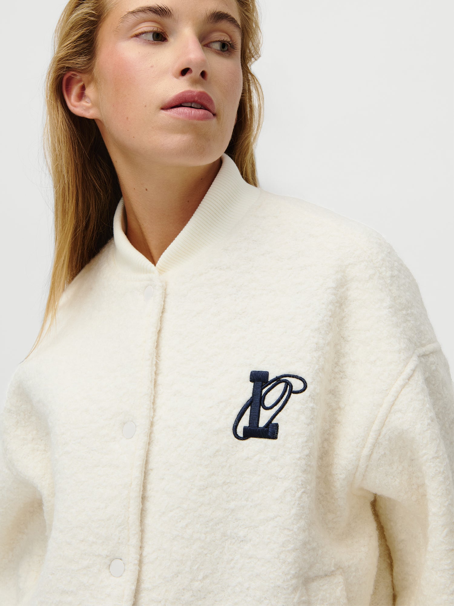 Eine Person, die die LeGer x Oh April - Blanca Jacket von LeGer by Lena Gercke trägt - eine strukturierte weiße Button-up-Jacke mit langen Ärmeln, geripptem Kragen und einem dunkelblauen, aufgestickten "L" auf der linken Brust - blickt leicht nach links vor einem einfarbigen Hintergrund.