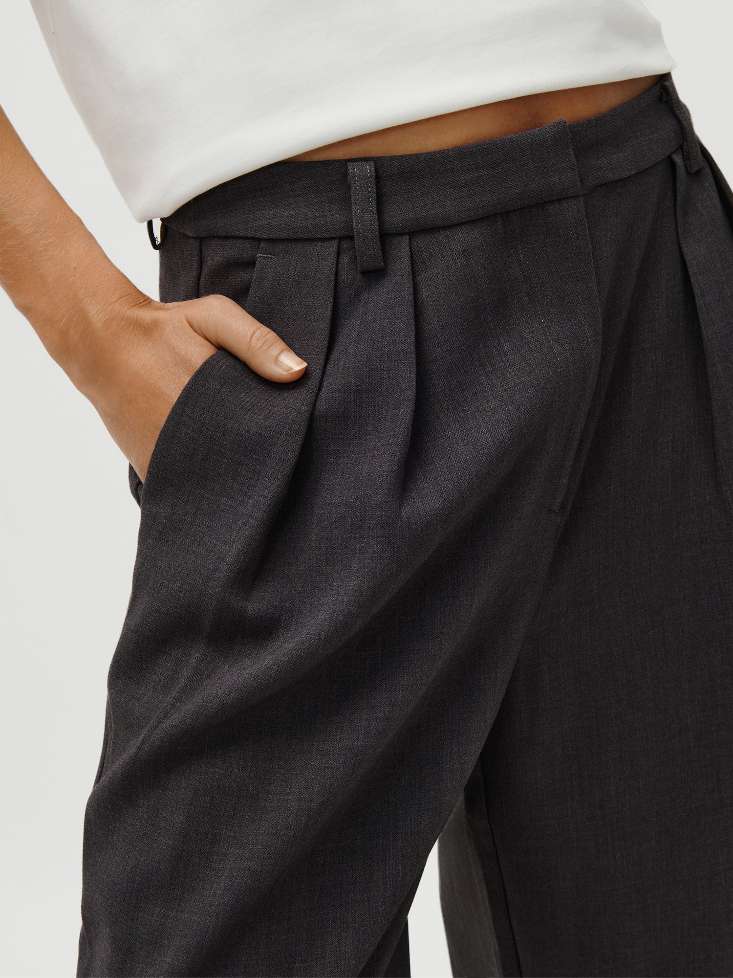 Eine Person steht mit einer Hand in der Tasche und trägt eine dunkle, plissierte LeGer x Oh April - Franja Pants von LeGer by Lena Gercke und ein weißes Top. Das Model ist 1,73 m groß und trägt Größe 36.