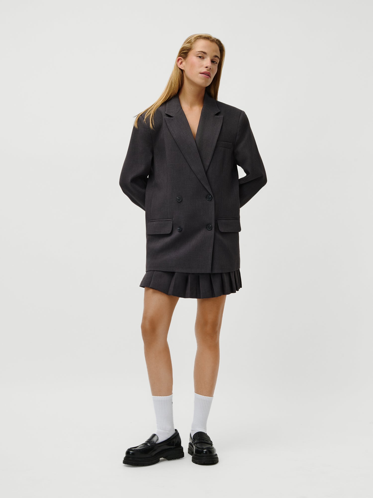 Eine Frau mit langem glattem Haar und neutralem Gesichtsausdruck trägt den LeGer x Oh April - Franja Blazer von LeGer by Lena Gercke, gestylt im Oversized-Look mit einem passenden Faltenminirock, schwarzen Loafers und weißen Socken vor einem schlichten weißen Hintergrund.