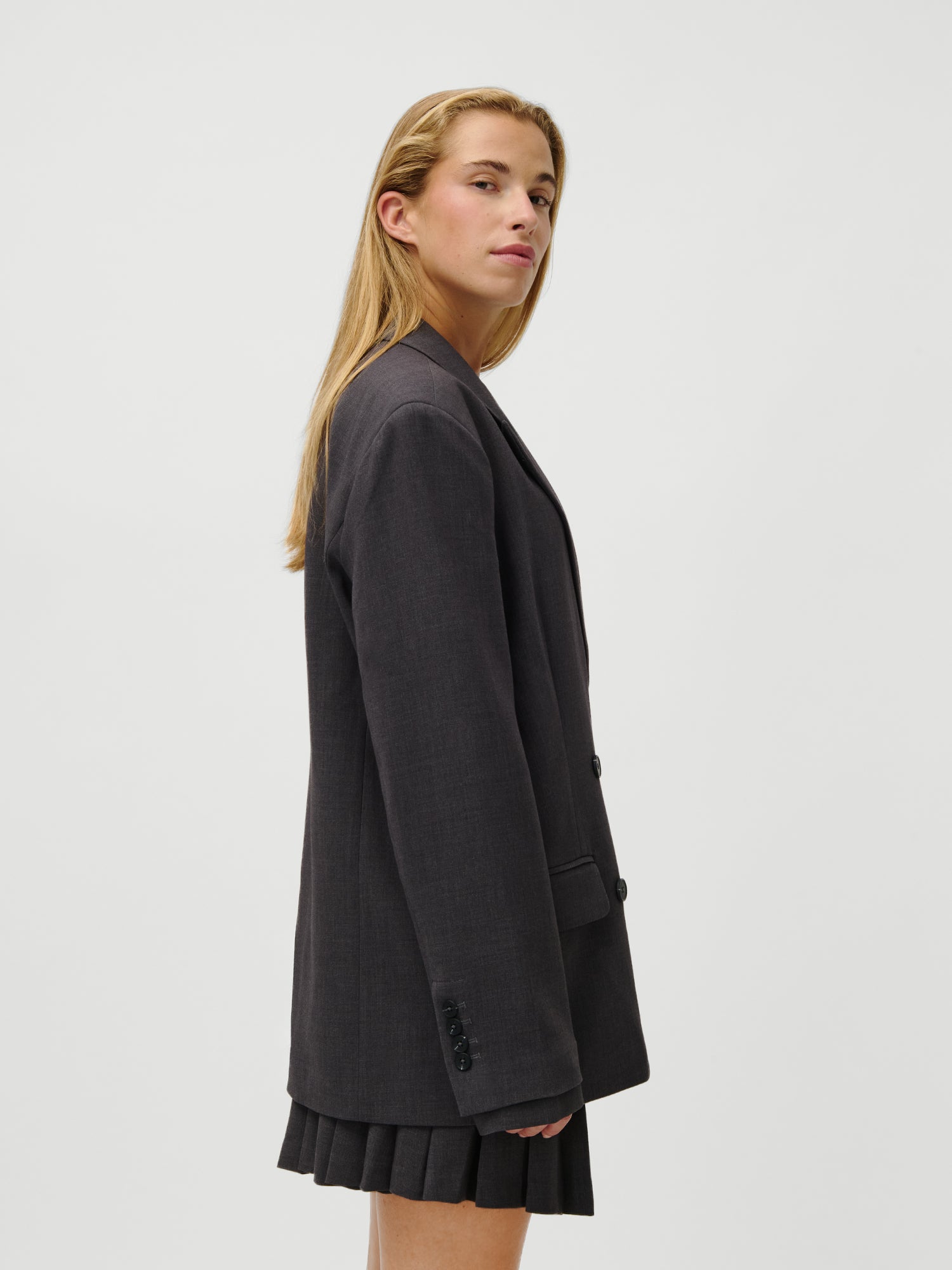 Eine Frau mit langen blonden Haaren steht seitlich, trägt den LeGer x Oh April - Franja Blazer von LeGer by Lena Gercke in einer lässigen Passform und einen schwarzen Faltenrock und schaut selbstbewusst in die Kamera vor einem schlichten weißen Hintergrund.