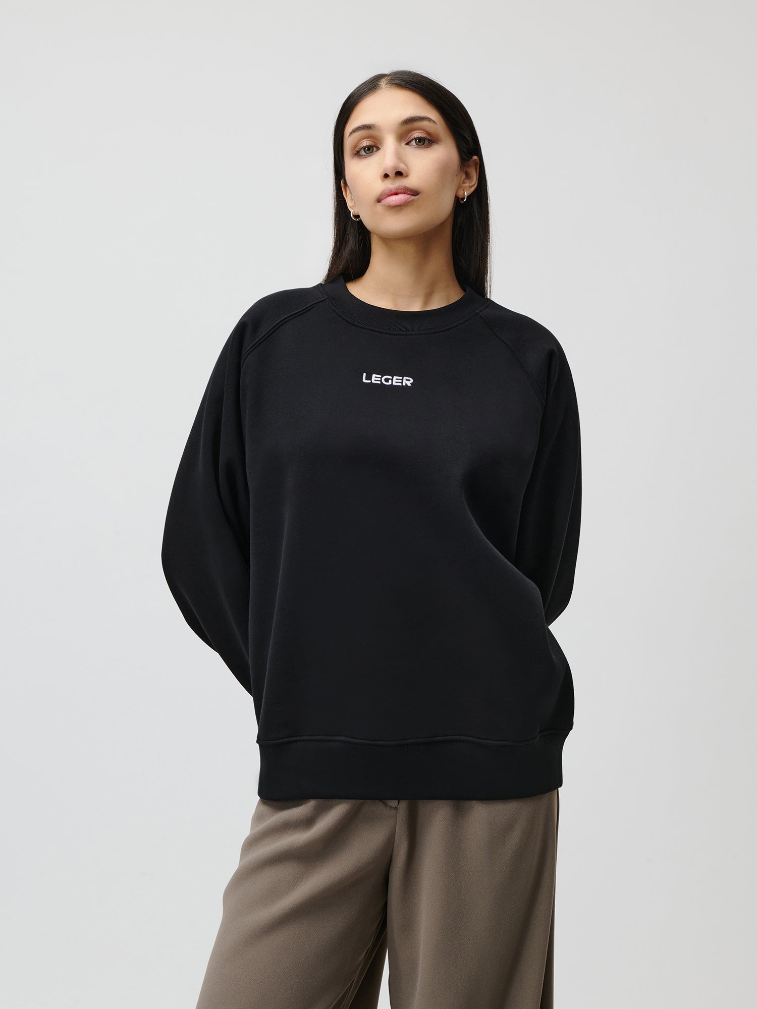 Eine Frau steht vor einem schlichten hellen Hintergrund. Sie trägt den Emma-Pullover von LeGer by Lena Gercke mit dem weißen Aufdruck „LECER“ auf der Vorderseite und eine taupefarbene Hose mit weitem Bein und hat die Hände für einen entspannten Sitz hinter dem Rücken verschränkt.