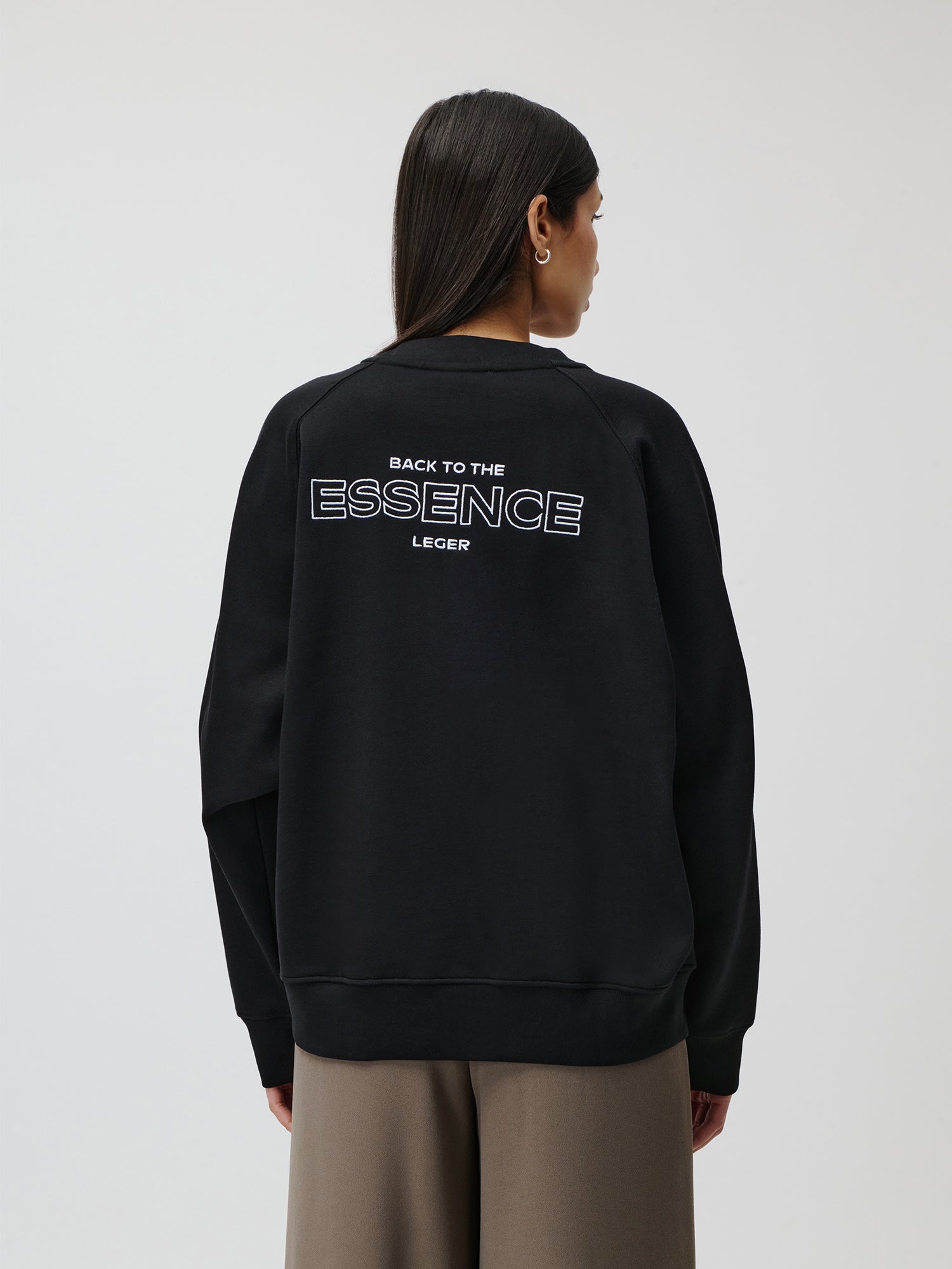 Eine Person mit langen dunklen Haaren, von hinten gezeigt, trägt den weiten schwarzen Emma Sweater von LeGer by Lena Gercke mit „BACK TO THE ESSENCE LEGER“ auf der Rückseite, kombiniert mit einer weiten braunen Hose und einer regulären Länge vor einem schlichten Hintergrund.