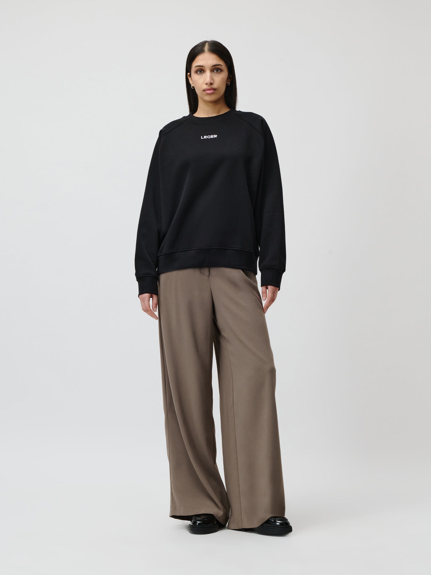 Eine Frau steht vor einem schlichten Hintergrund und trägt einen langärmeligen, lockeren schwarzen Emma-Pullover von LeGer by Lena Gercke in normaler Passform und Länge, eine weite taupefarbene Hose und schwarze Schuhe. Sie hat glattes dunkles Haar und einen neutralen Gesichtsausdruck.