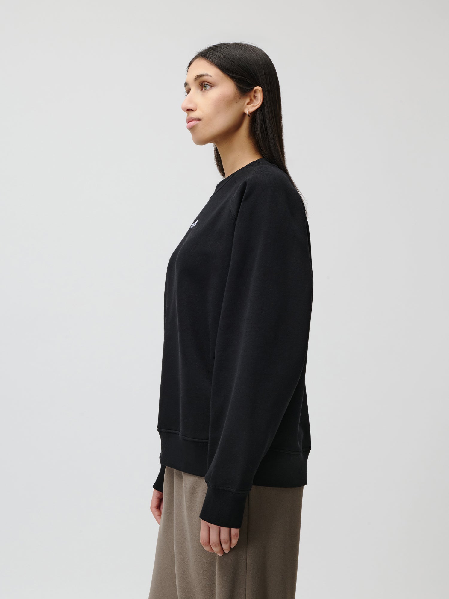 Eine Frau mit langem, dunklem Haar steht im Profil vor einem schlichten, hellen Hintergrund und trägt den Emma-Pullover von LeGer by Lena Gercke und taupefarbene Hosen mit weitem Bein.