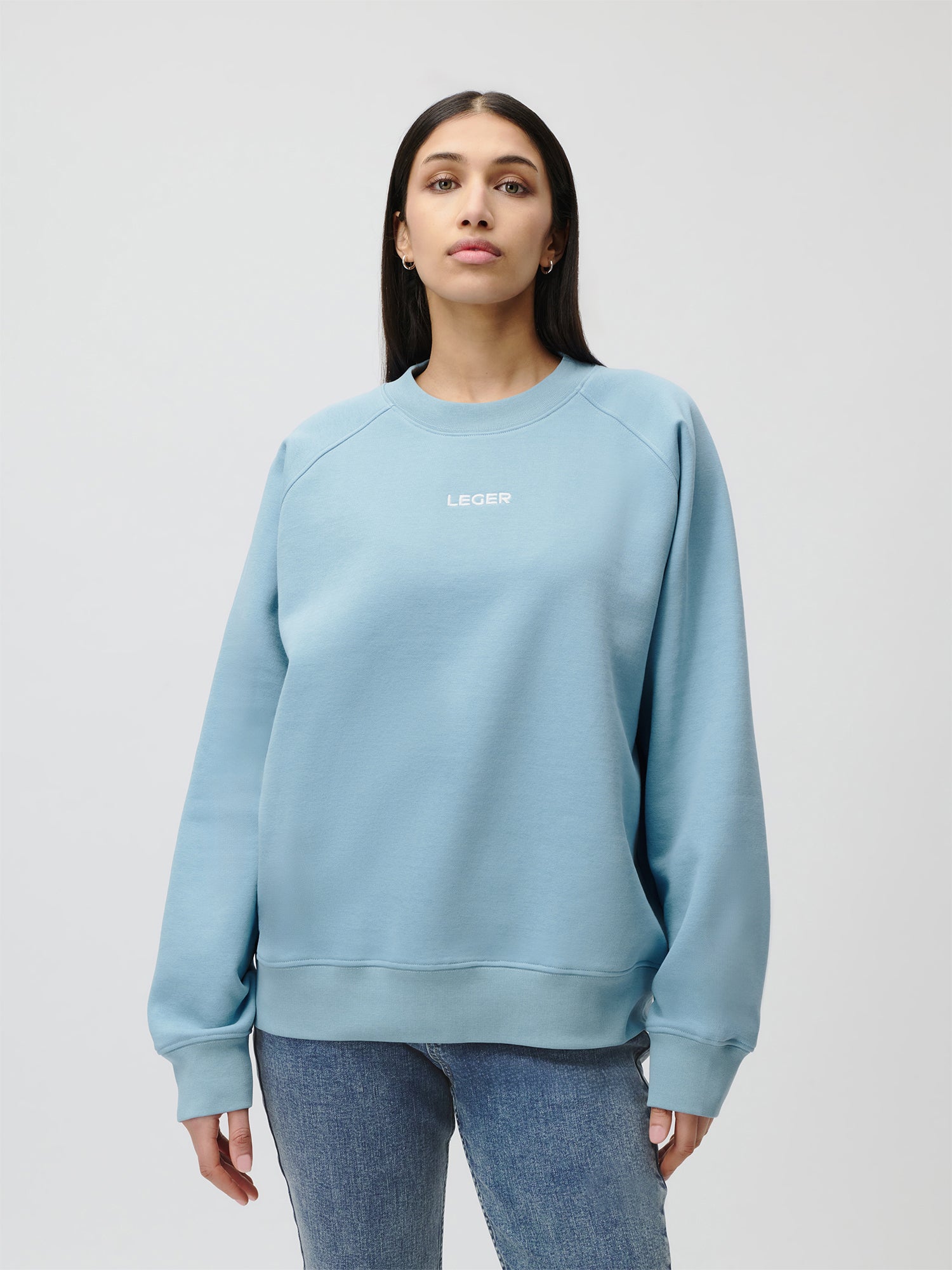Eine Frau blickt nach vorne und trägt den Emma Sweater von LeGer by Lena Gercke - ein hellblaues, normal langes Sweatshirt mit dem Schriftzug "LEGER" auf der Brust und langen Ärmeln, das mit einer blauen Jeans kombiniert wird. Der Hintergrund ist einfarbig und hell.