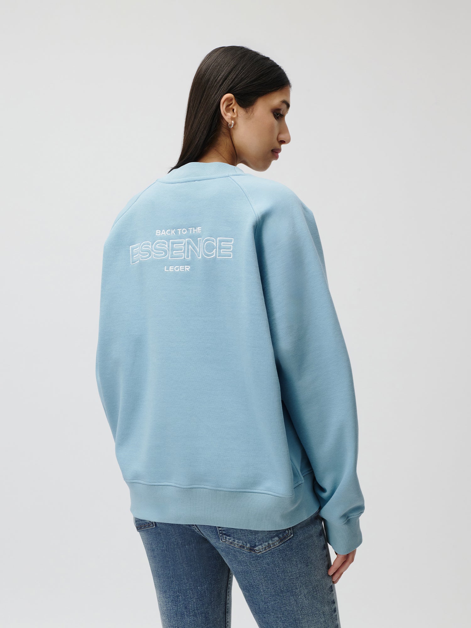 Eine Person mit langen dunklen Haaren trägt den Emma Sweater von LeGer by Lena Gercke in Hellblau mit dem Schriftzug "BACK TO THE ESSENCE LEGER" auf dem Rücken, gepaart mit einer blauen Jeans, vor einem schlichten hellen Hintergrund für einen Schrank-Look.