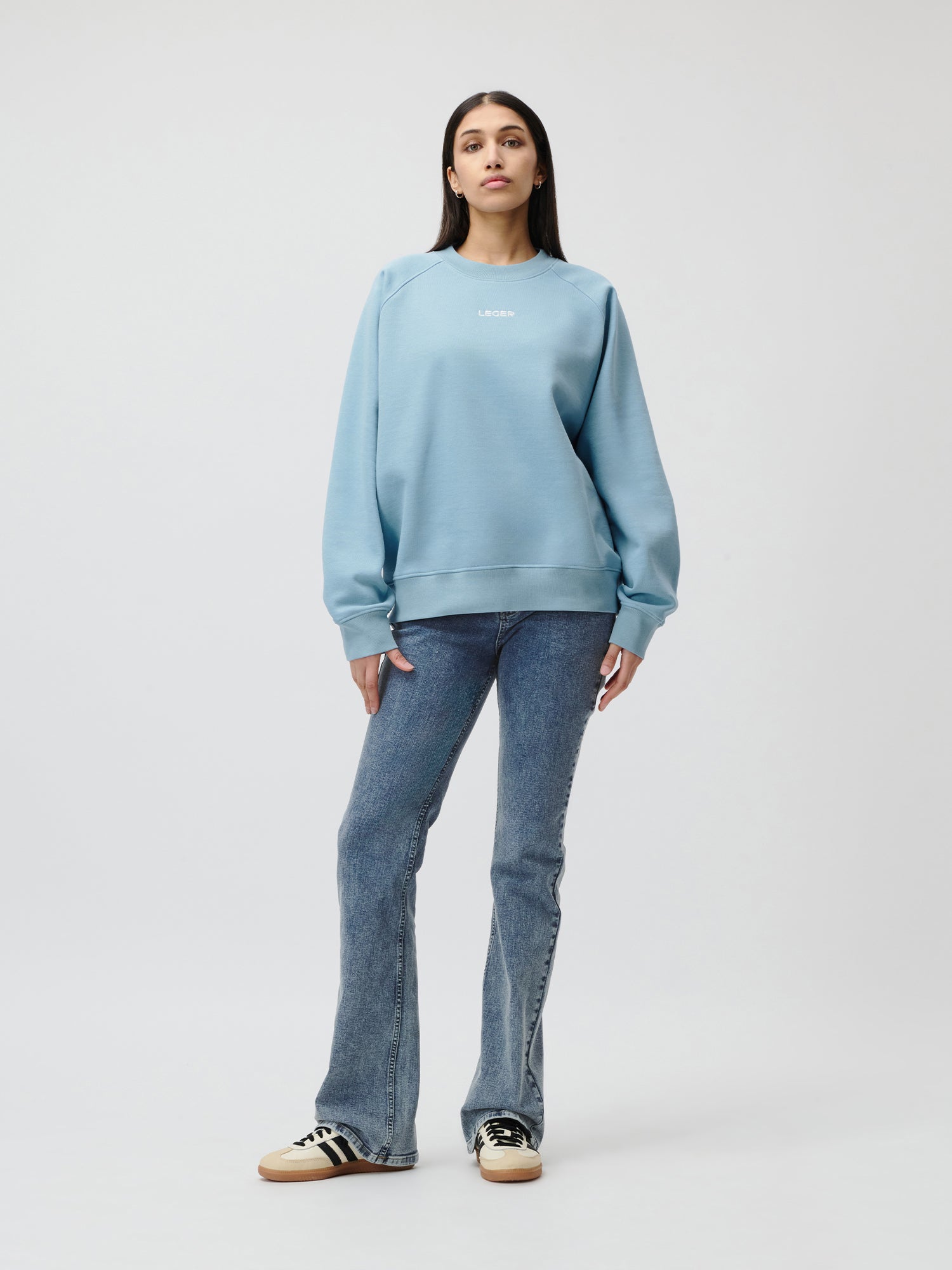 Eine Frau steht vor einem weißen Hintergrund und trägt den hellblauen Emma Sweater von LeGer by Lena Gercke, blaue Schlagjeans und weiße Turnschuhe mit schwarzen Streifen. Ihr Blick ist nach vorne gerichtet, ihre Arme sind entspannt und sie sieht lässig aus.