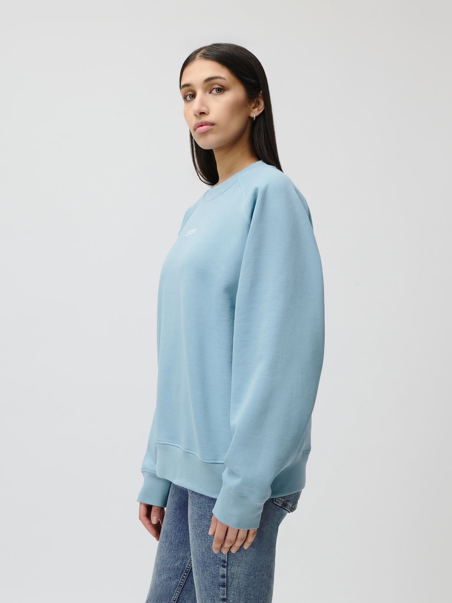 Eine Frau mit glattem dunklem Haar steht seitlich und schaut in die Kamera. Sie trägt den Emma Sweater von LeGer by Lena Gercke in Hellblau und Blue Jeans vor einem schlichten weißen Hintergrund.