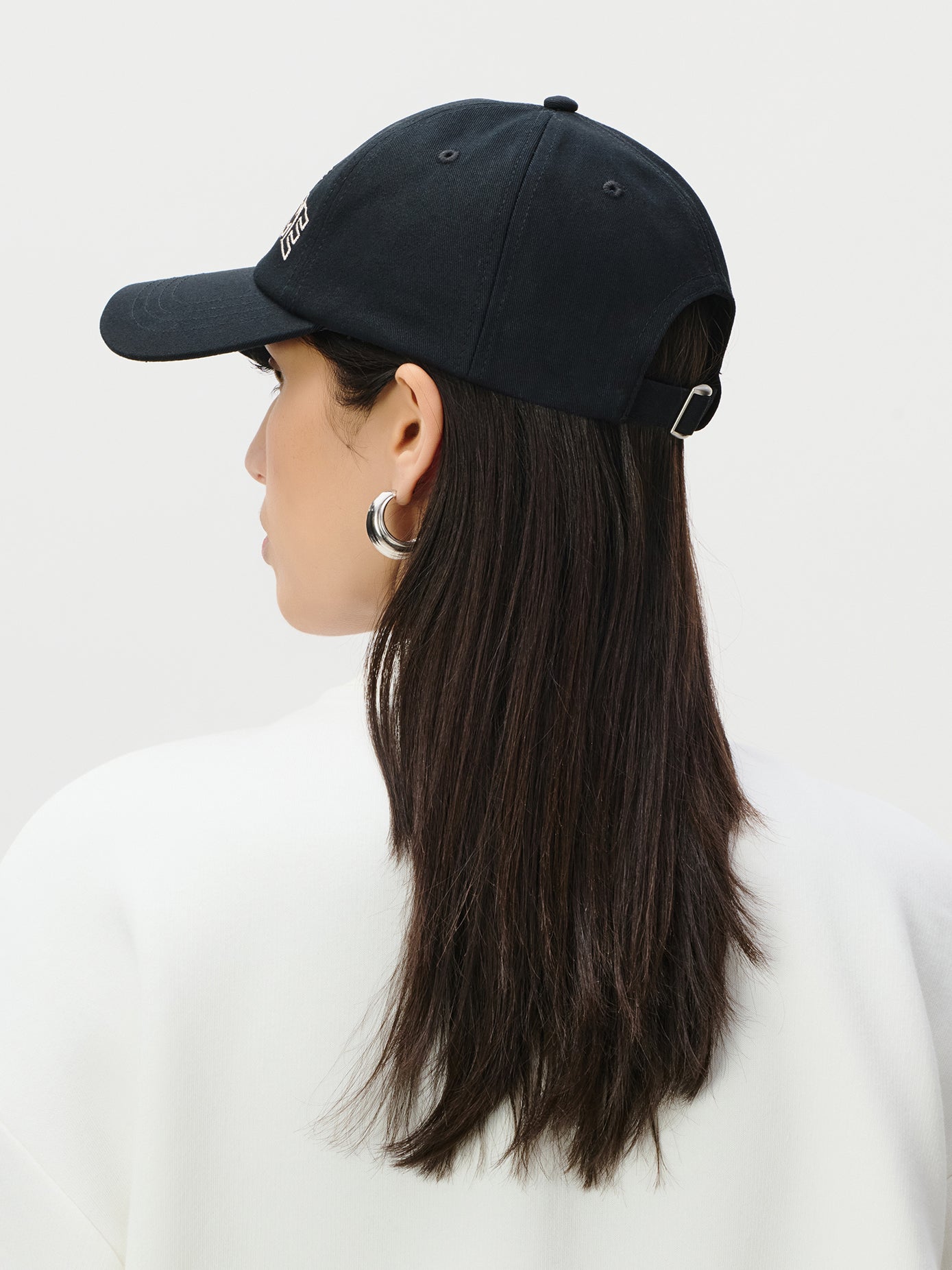 Eine Person mit glattem, dunkelbraunem Haar trägt die LeGer by Lena Gercke Iris Cap in Schwarz und silberne Ohrringe, von der Kamera abgewandt. Sie hat ein weißes Oberteil vor einem schlichten hellen Hintergrund an.