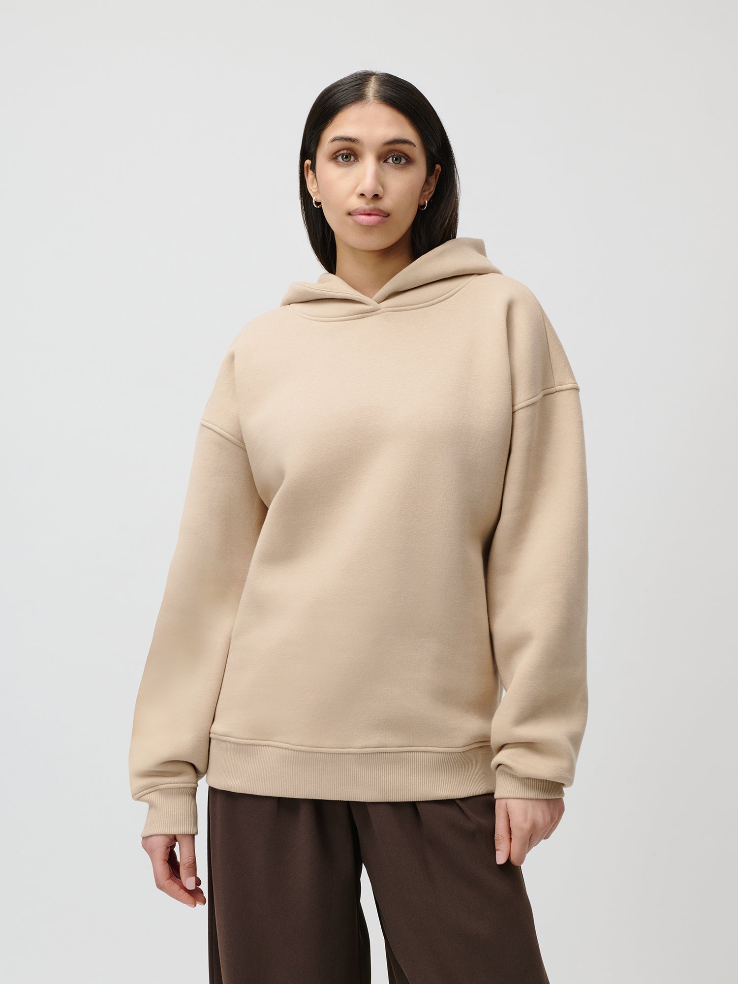 Eine Frau mit langen dunklen Haaren trägt den beigen Aylin Hoodie von LeGer by Lena Gercke in Größe 36 und eine braune Hose, steht vor einem schlichten hellgrauen Hintergrund und blickt mit neutralem Gesichtsausdruck in die Kamera.