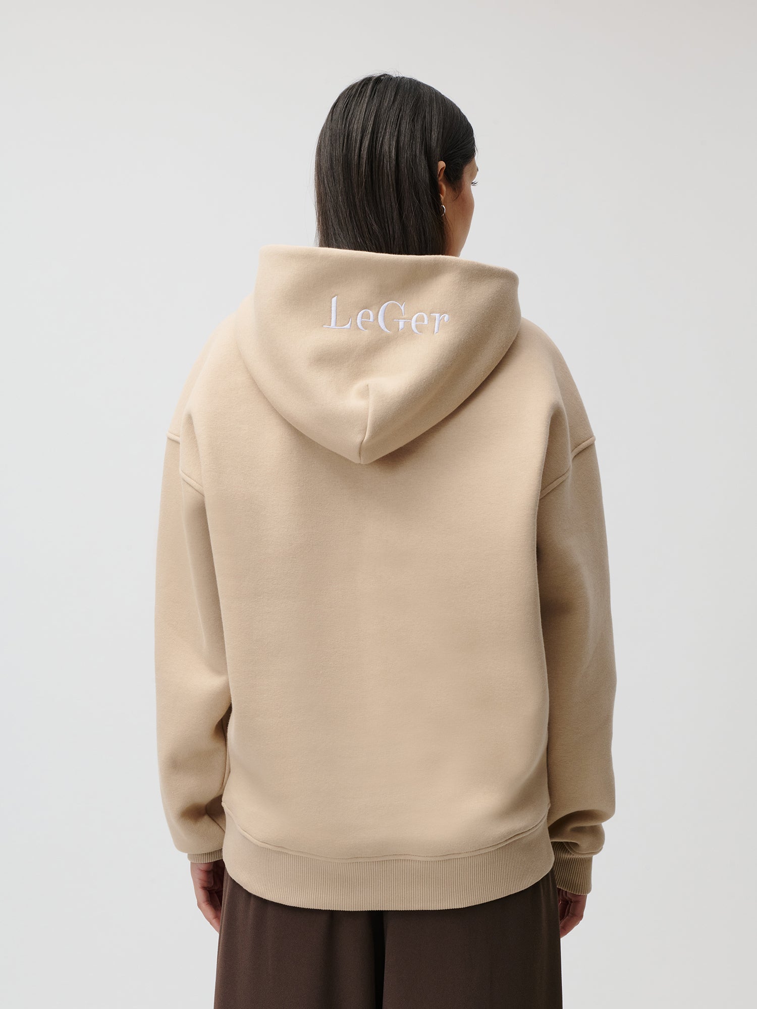 Eine Person mit glattem, dunklem Haar blickt abgewandt und trägt einen beigen Aylin Hoodie in Größe 36 von LeGer by Lena Gercke mit „LeGer“ in Weiß auf der Kapuze. Der Hintergrund ist schlicht und hell.