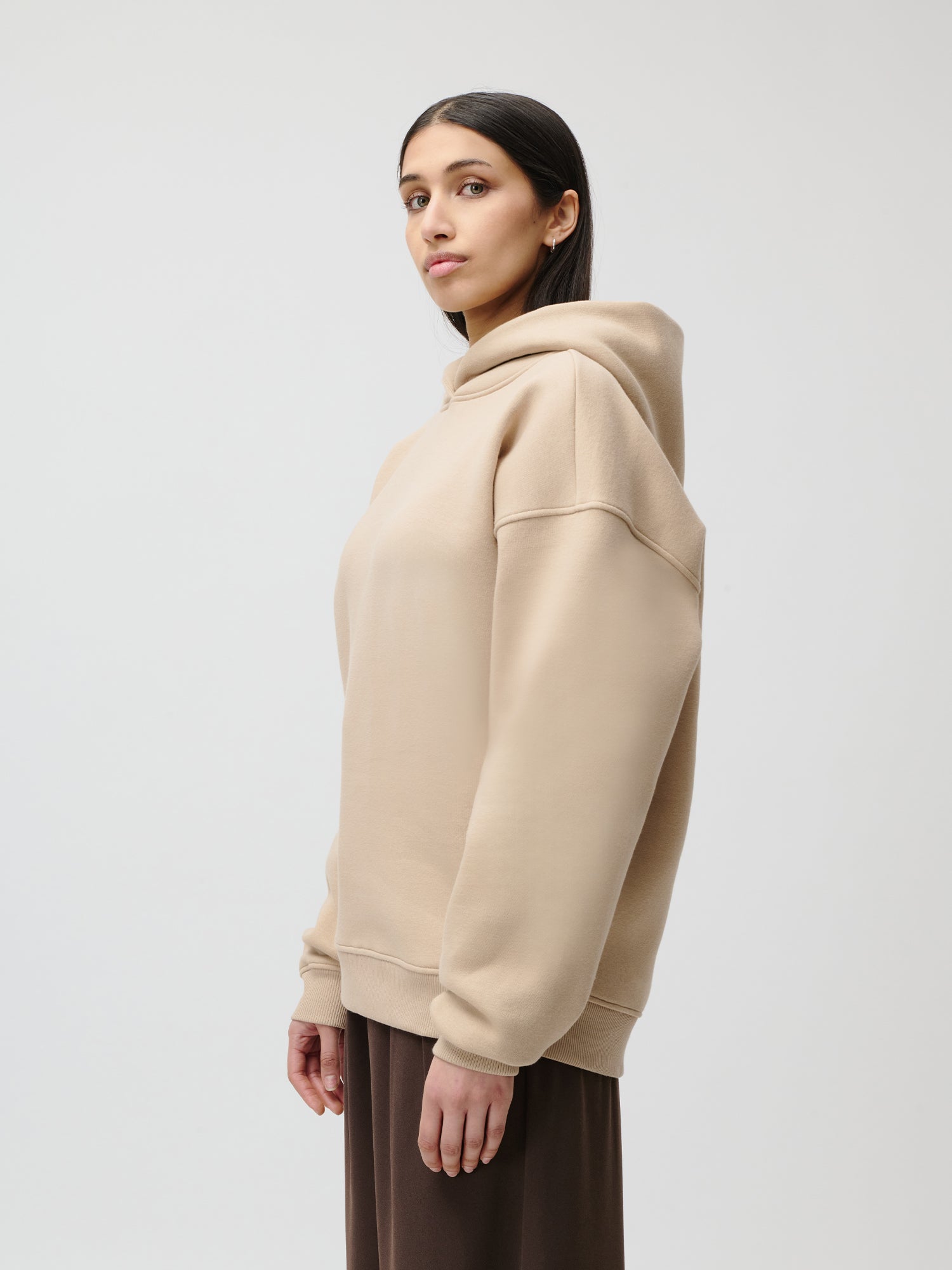 Eine Frau mit glattem, dunklem Haar, die einen beigen Aylin Hoodie von LeGer by Lena Gercke und einen braunen Rock in Größe 36 trägt, steht seitlich vor einem schlichten, hellen Hintergrund und blickt in die Kamera.
