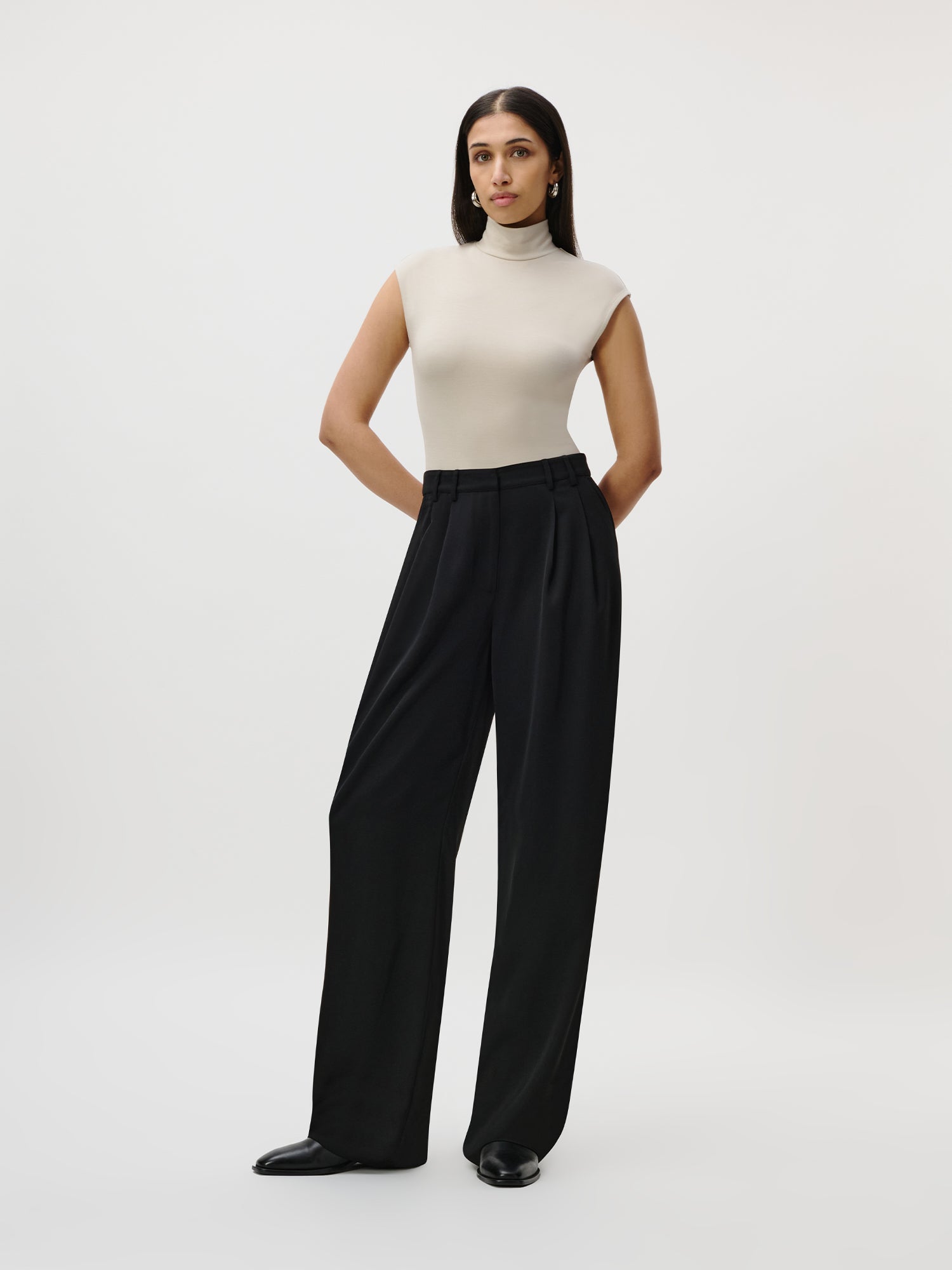Eine Frau mit glattem, dunklem Haar steht vor einem schlichten, hellen Hintergrund. Sie trägt einen ärmellosen, beigen Rollkragenpullover, der in die Kora Pants Tall von LeGer by Lena Gercke gesteckt ist, die einen hoch taillierten Schnitt und eine dezente Satinierung aufweist. Ihre Hände sind hinter dem Rücken verschränkt.