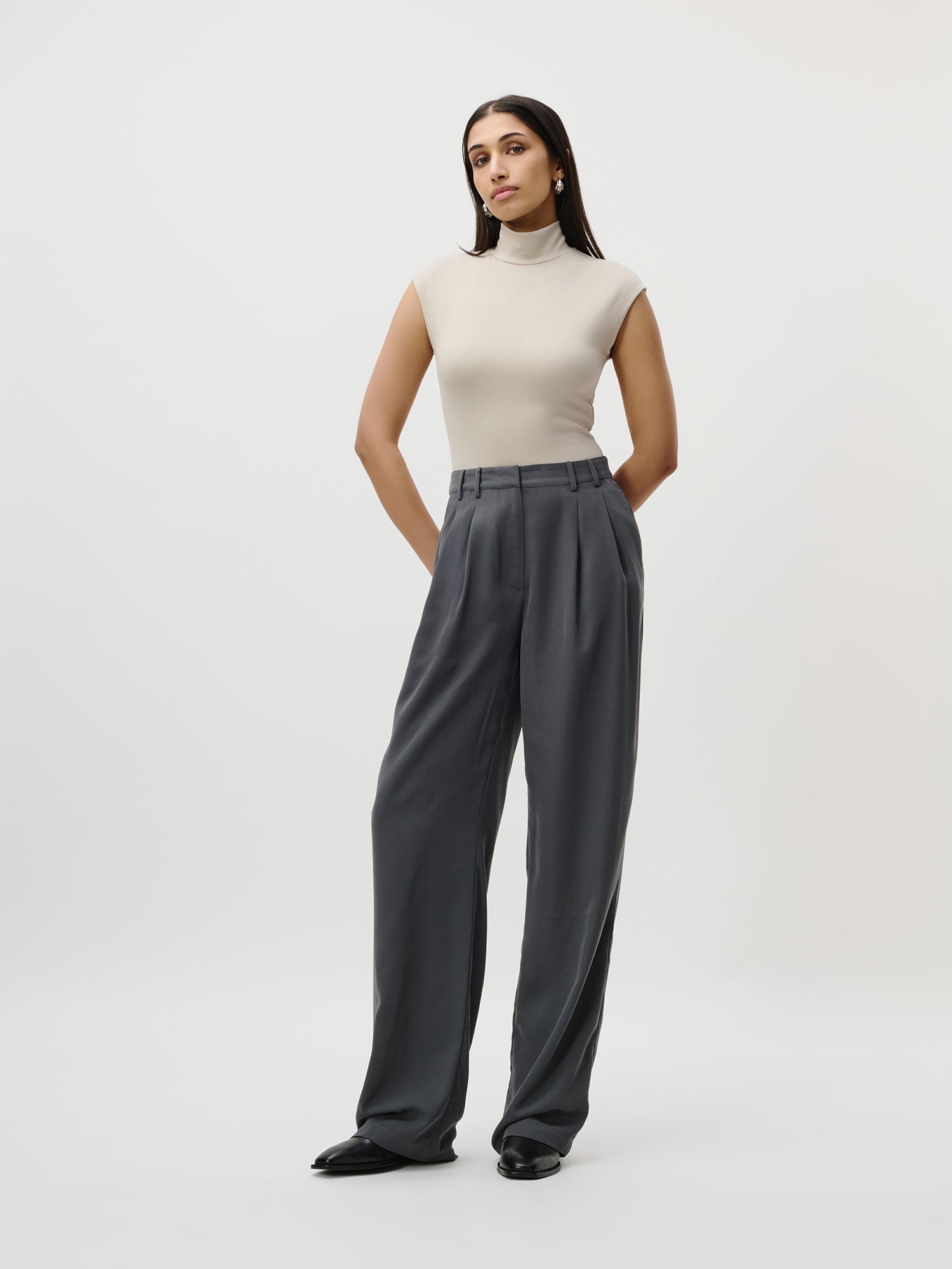 Eine Frau mit glattem dunklem Haar posiert mit den Händen in der Tasche vor einem schlichten weißen Hintergrund. Sie trägt die Kora Pants Tall von LeGer by Lena Gercke, eine graue Hose mit hoher Taille und weitem Bein, die mit einem ärmellosen beigen Rollkragenpullover kombiniert wird und einen eleganten Look ergibt.
