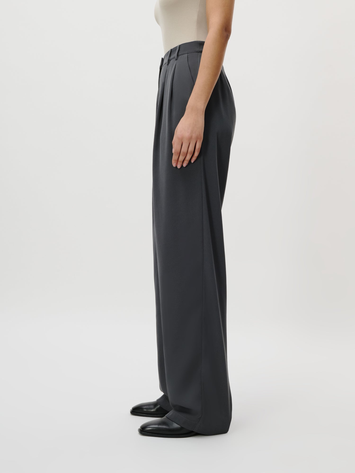 Eine Person in Größe 36 LeGer by Lena Gercke Kora Pants Tall - mit hoher Taille, weitem Bein, dunkelgrau - wird im Seitenprofil gezeigt, die ein umgeschlagenes beigefarbenes ärmelloses Oberteil und schwarze Schuhe vor einem schlichten weißen Hintergrund trägt.