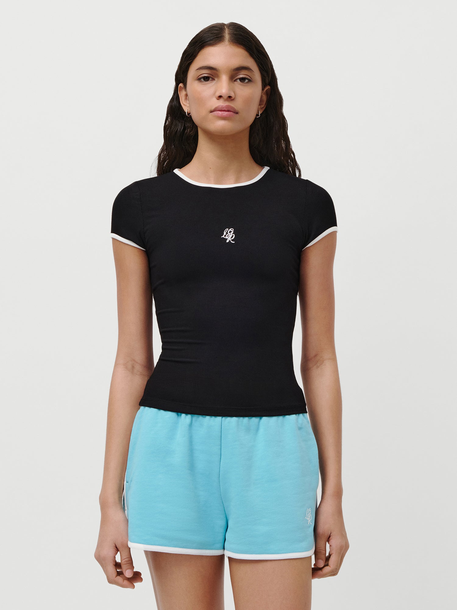 Eine Frau mit langen dunklen Haaren trägt das LeGer by Lena Gercke Elmira T-Shirt, ein schmal geschnittenes schwarzes T-Shirt mit weißem Rand und einem kleinen weißen Brustlogo, gepaart mit hellblauen Sportshorts, vor einem schlichten weißen Hintergrund.