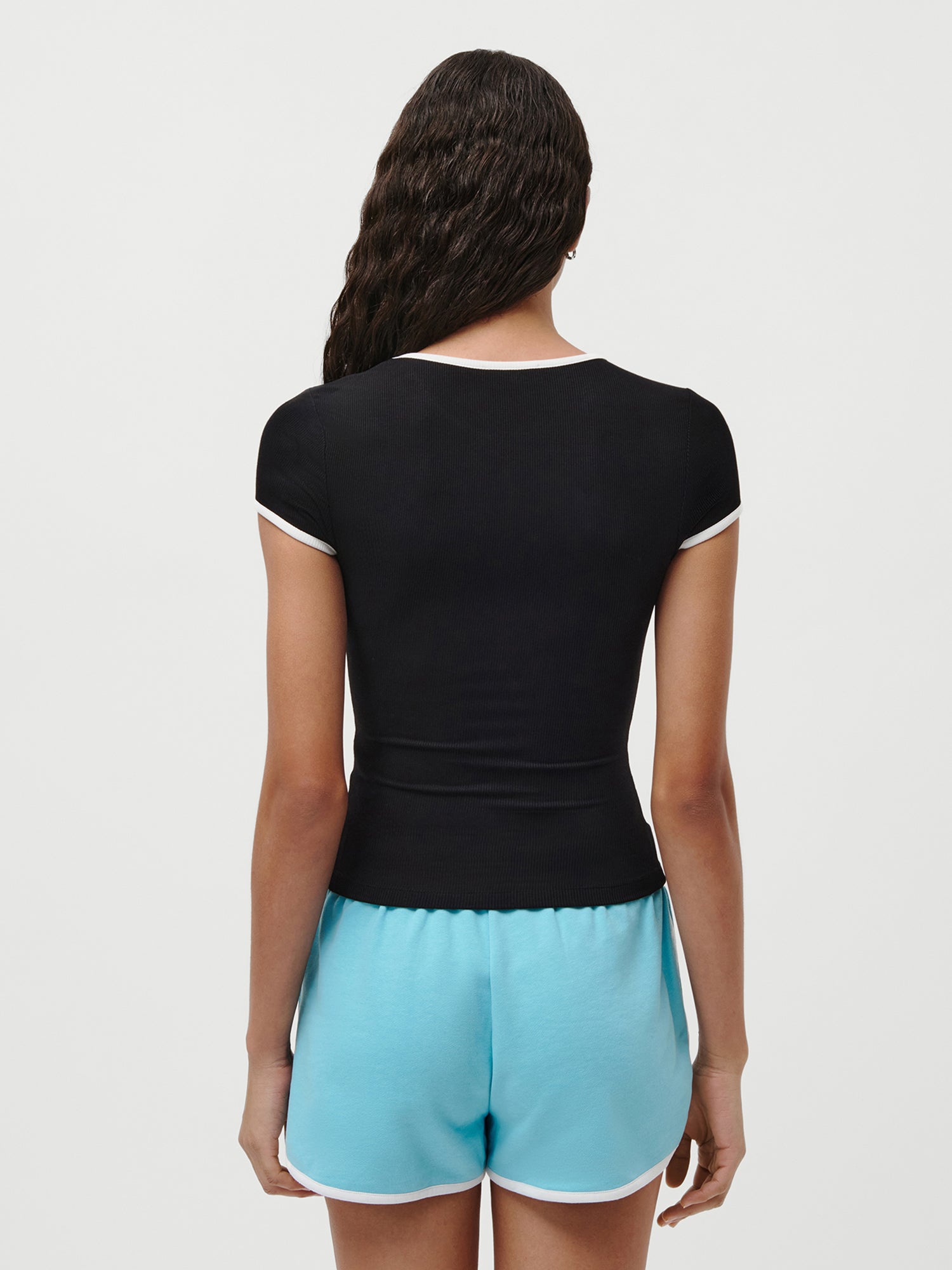 Eine Person mit langen, gewellten Haaren ist von hinten zu sehen. Sie trägt ein schwarzes Elmira-T-Shirt von LeGer by Lena Gercke mit weißem Saum und hellblaue Sportshorts mit weißem Bund vor einem schlichten weißen Hintergrund.