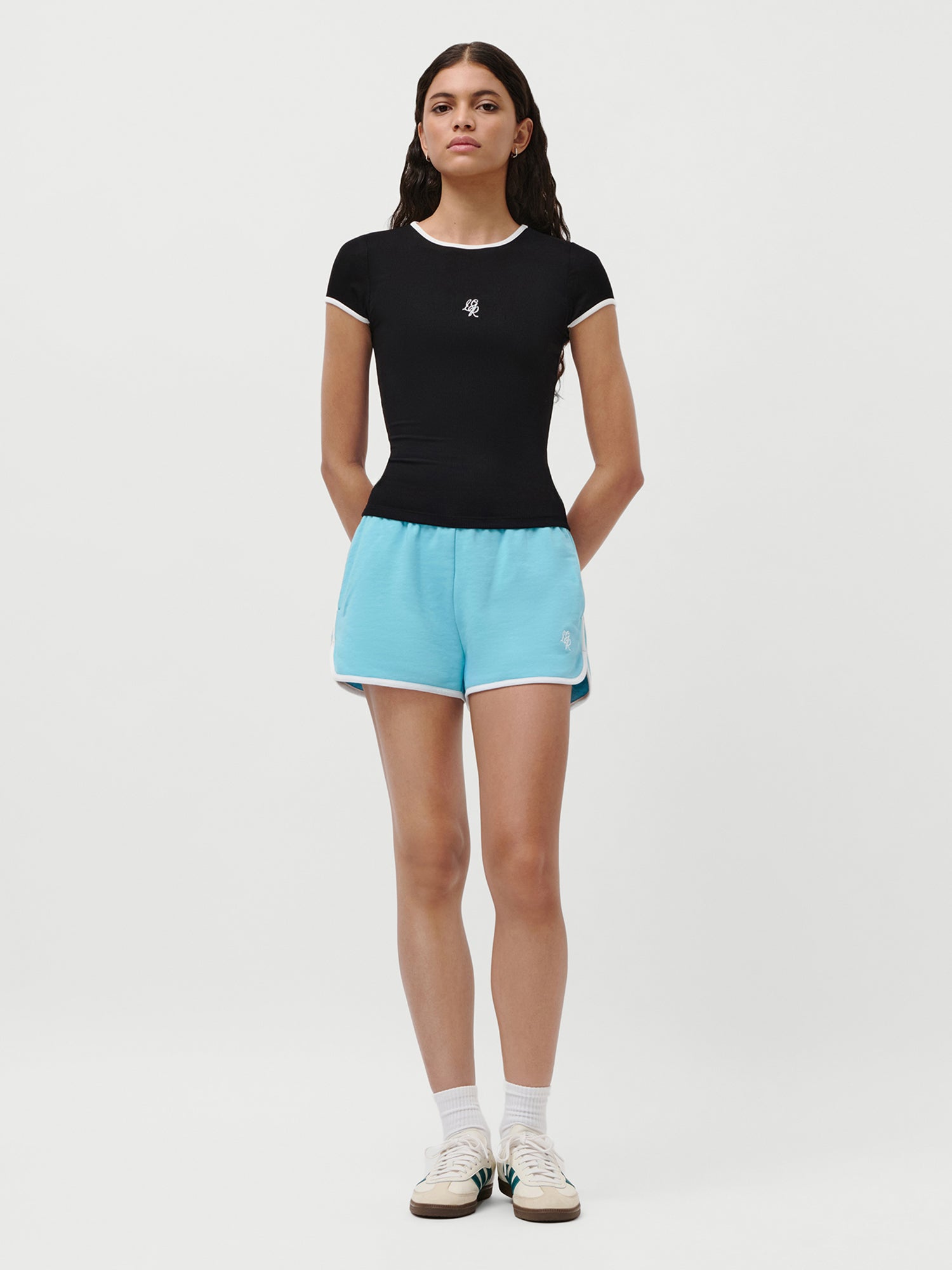 Eine Frau blickt nach vorne und trägt das Elmira T-Shirt von LeGer by Lena Gercke in Schwarz (schmale Passform, kurze Ärmel), hellblaue Sporthosen, weiße Socken und weiße Turnschuhe. Sie steht vor einem schlichten weißen Hintergrund und hat die Hände in den Taschen ihrer Shorts.