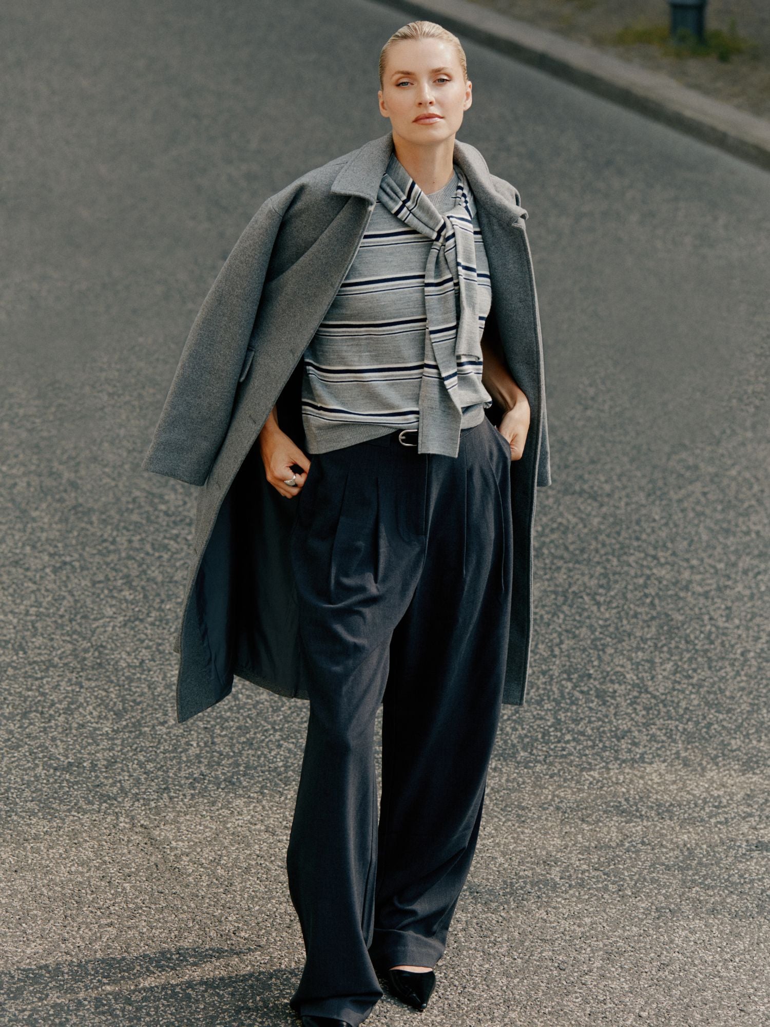 Eine Person steht auf einer Straße und trägt den Selma Pullover von LeGer by Lena Gercke in Größe 36, gestylt mit einer dunklen Hose mit weitem Bein und einem langen grauen Mantel, der über die Schultern gelegt ist.